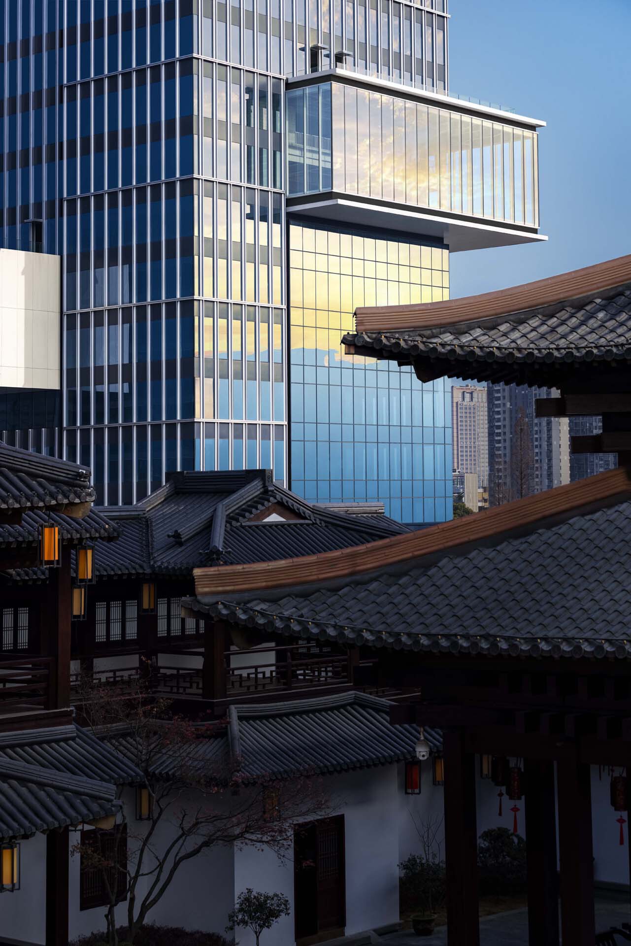 Glass tower with cantilevered volume rising behind traditional timber and tile roofed buildings at dusk