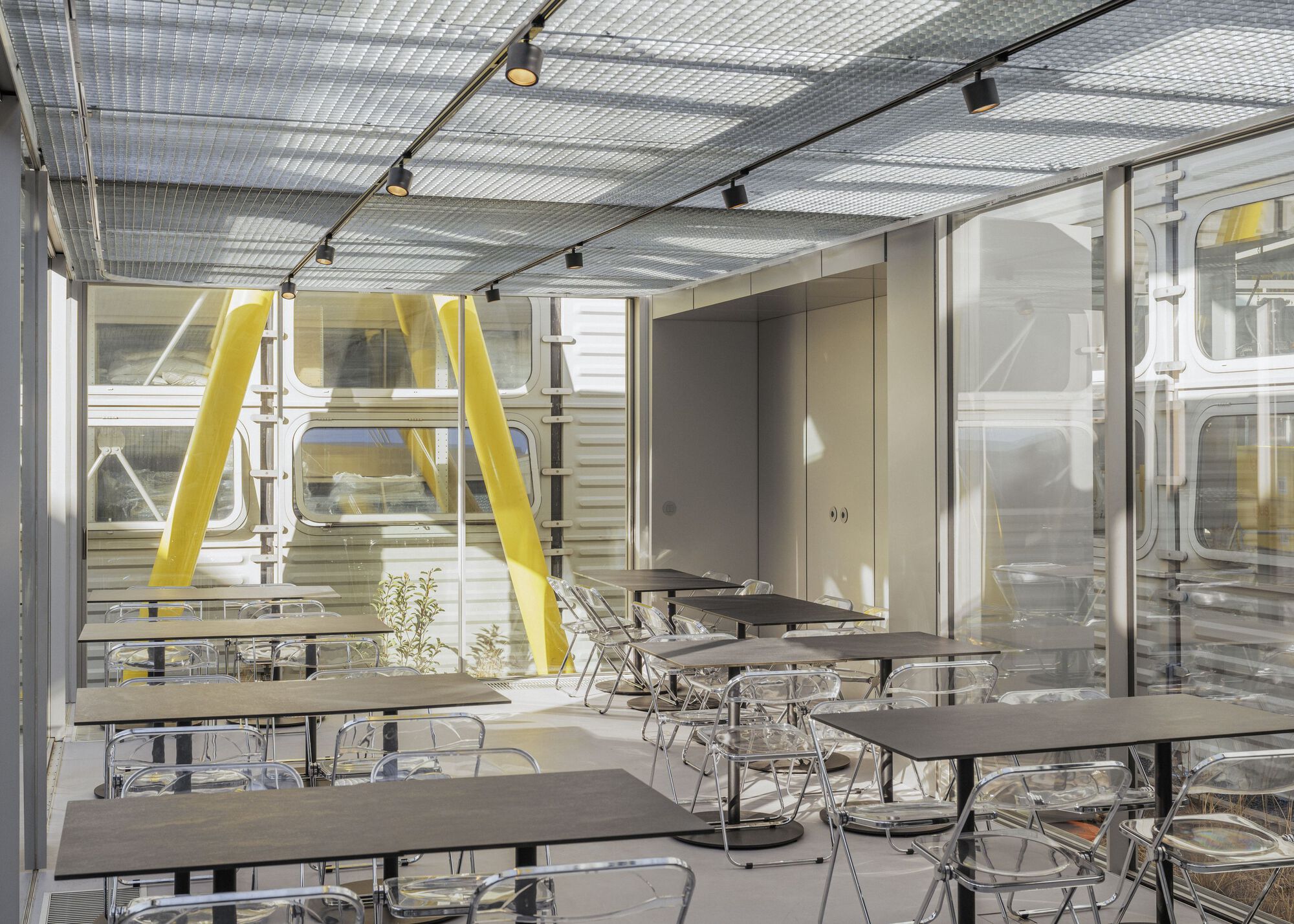 Interior dining area with translucent corrugated ceiling panels, track lighting, and yellow structural columns visible through glazing