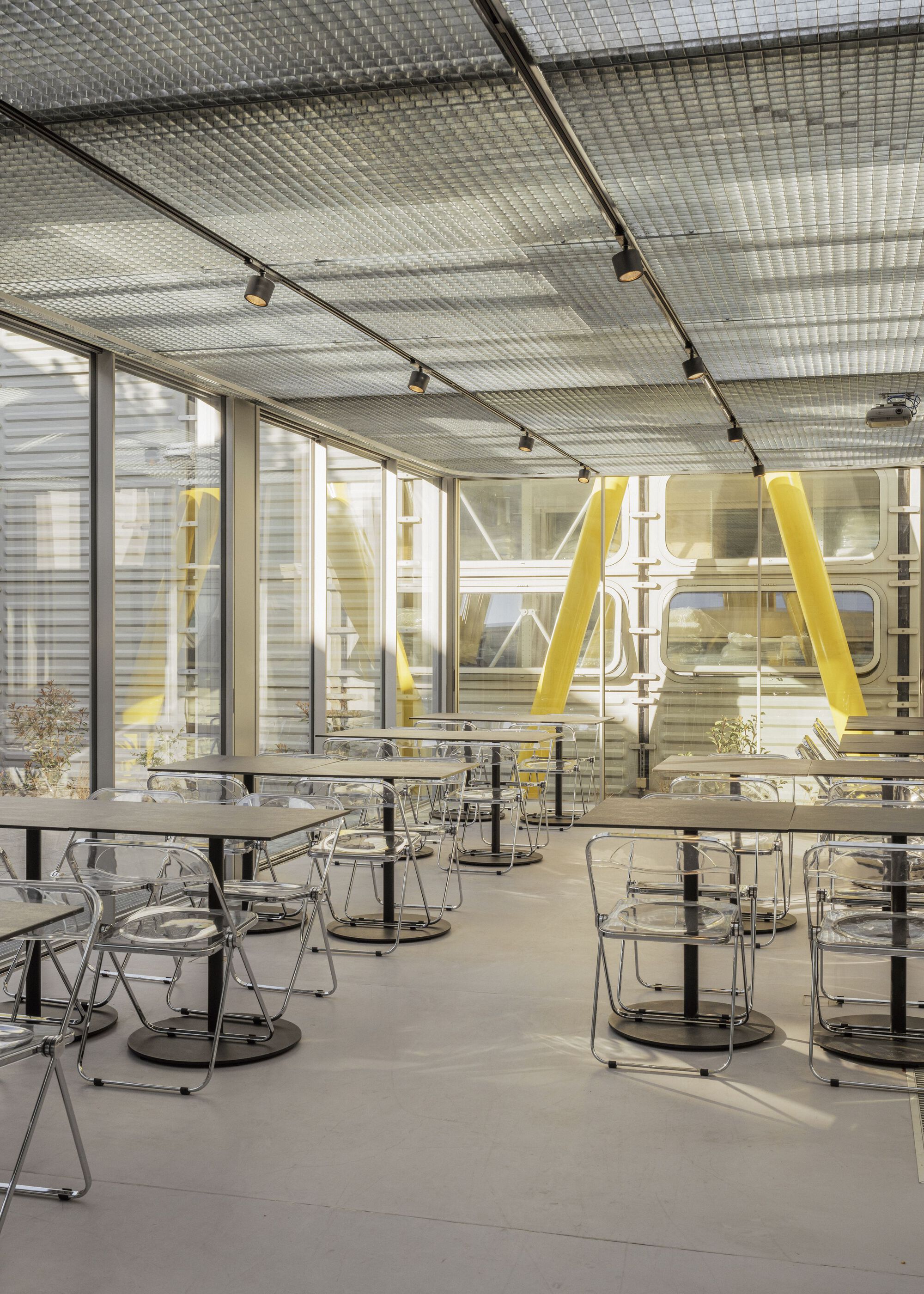 Dining space with mesh metal ceiling, transparent chairs, and yellow diagonal bracing seen through glass partition
