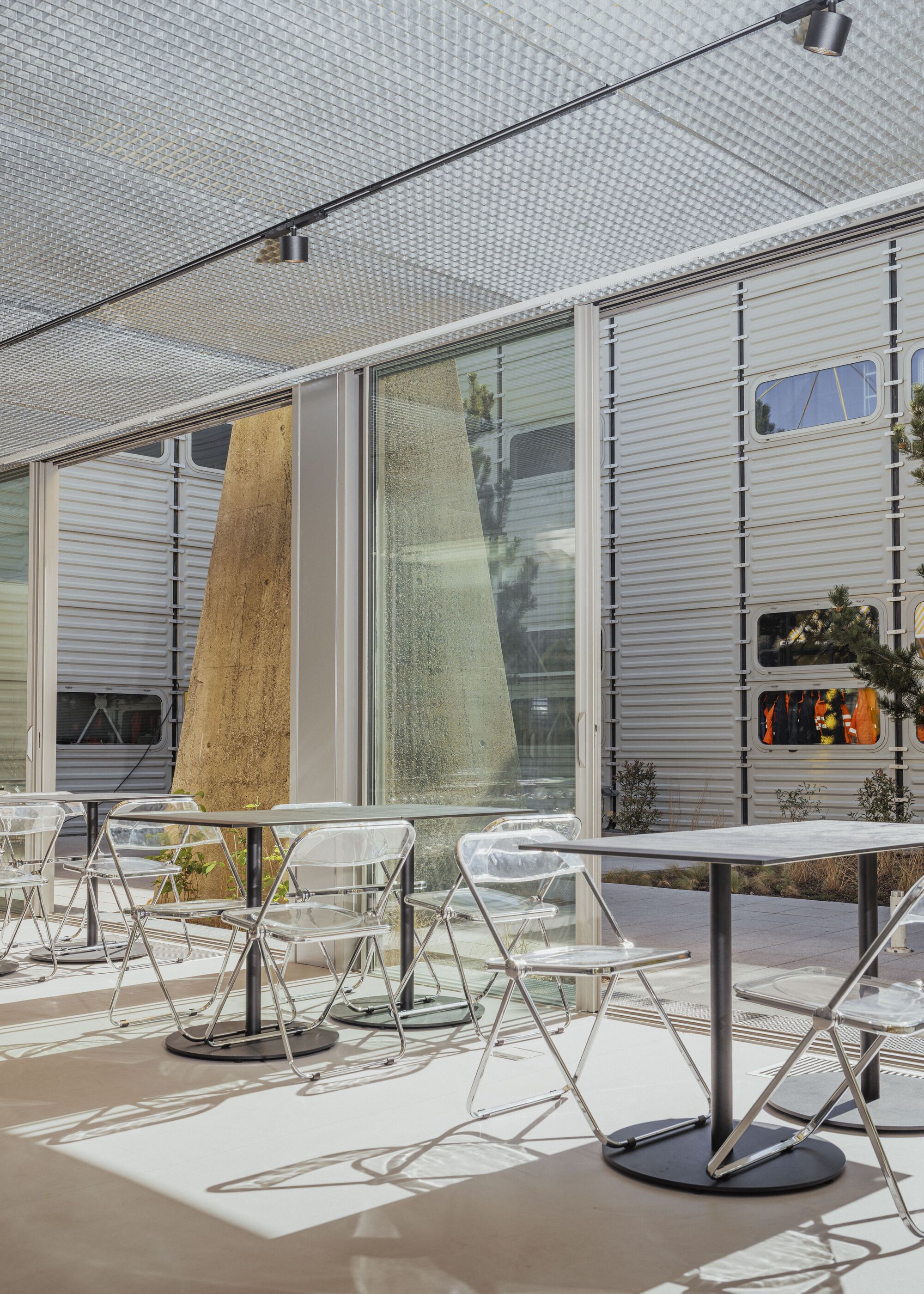 Outdoor seating terrace beneath perforated metal canopy with corrugated facade panels and planted beds in afternoon light