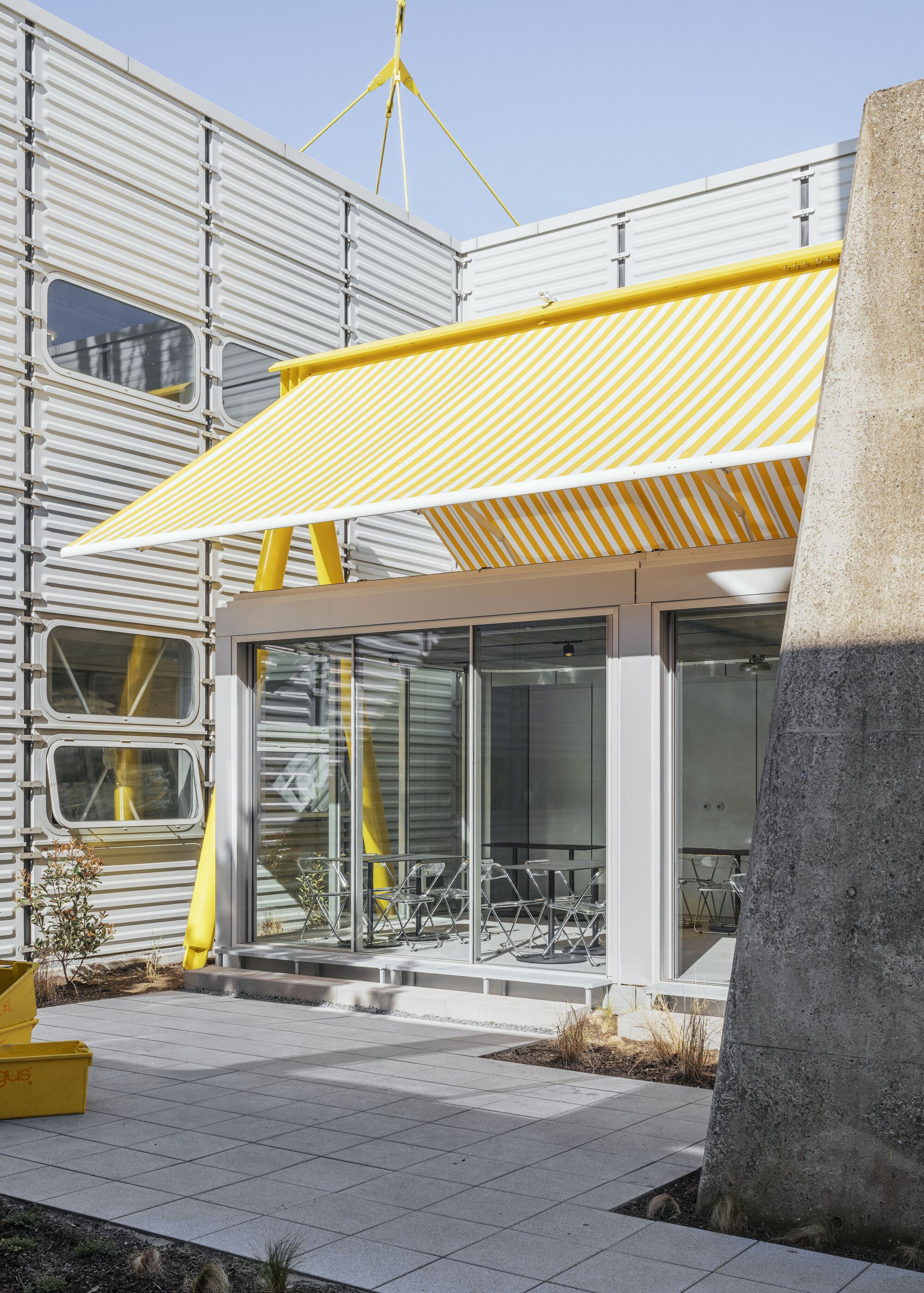 Covered entry threshold with yellow striped fabric canopy, concrete column, and corrugated metal facade under blue sky