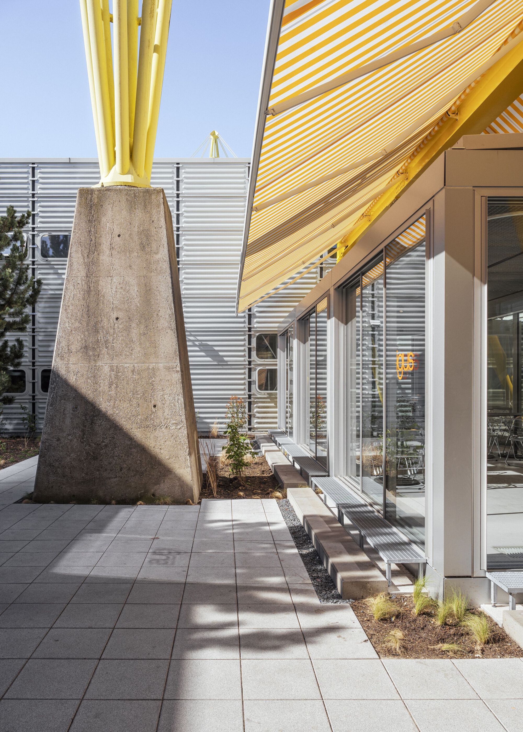Tapered concrete column supporting yellow striped canopy beside corrugated metal facade and paved courtyard