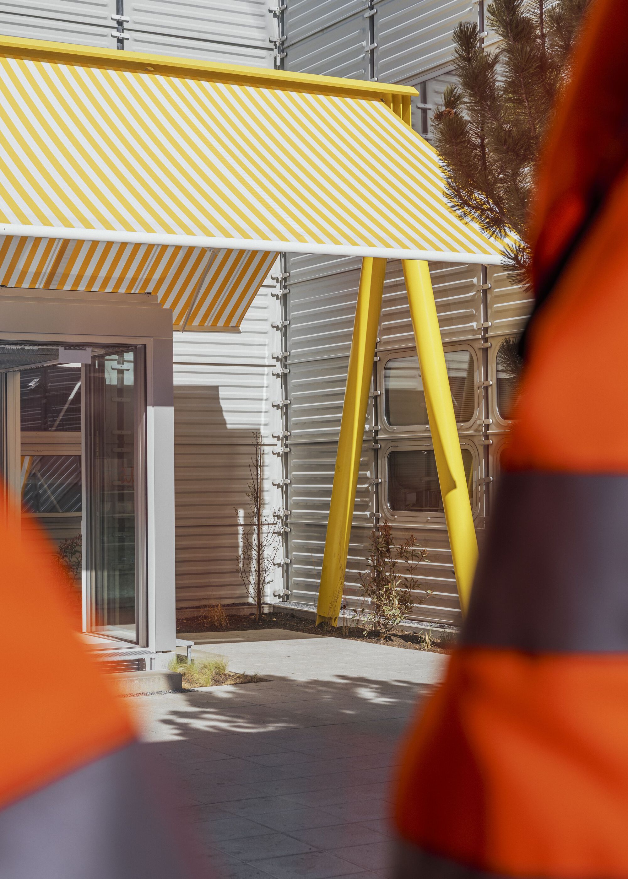 Courtyard view through orange bollards showing yellow canopy and planted beds along corrugated facade