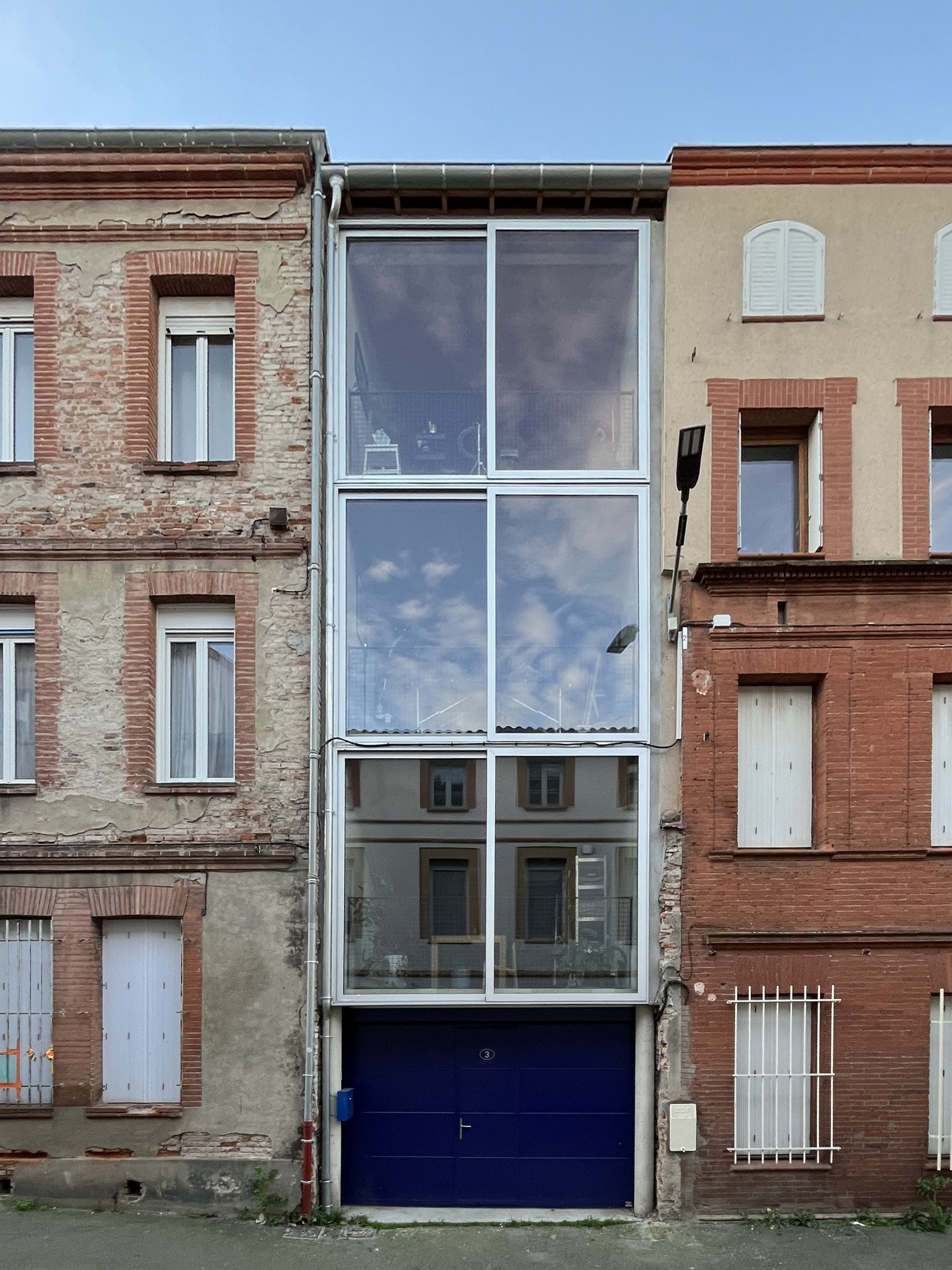 Narrow glass facade with white frames and a blue garage door inserted between weathered brick rowhouses