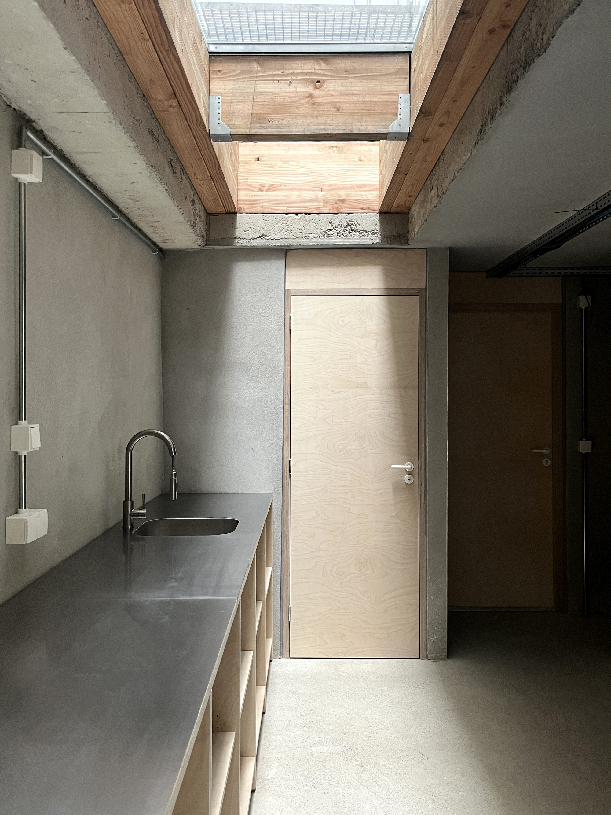 Concrete and timber kitchen corridor with rectangular skylight illuminating the light wood door