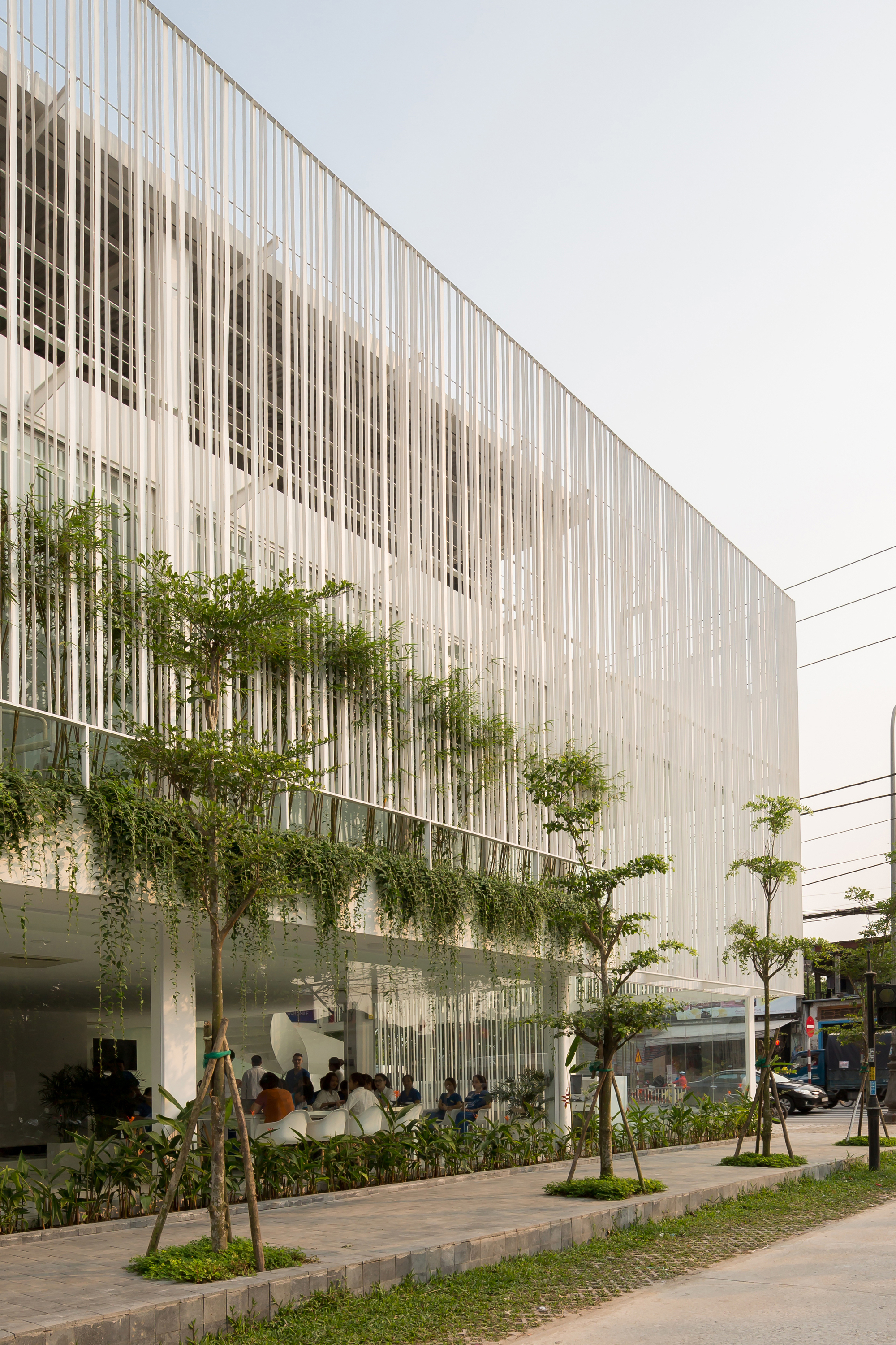 Exterior view showing vertical white louvers above a planted terrace with bamboo and pedestrians on the sidewalk