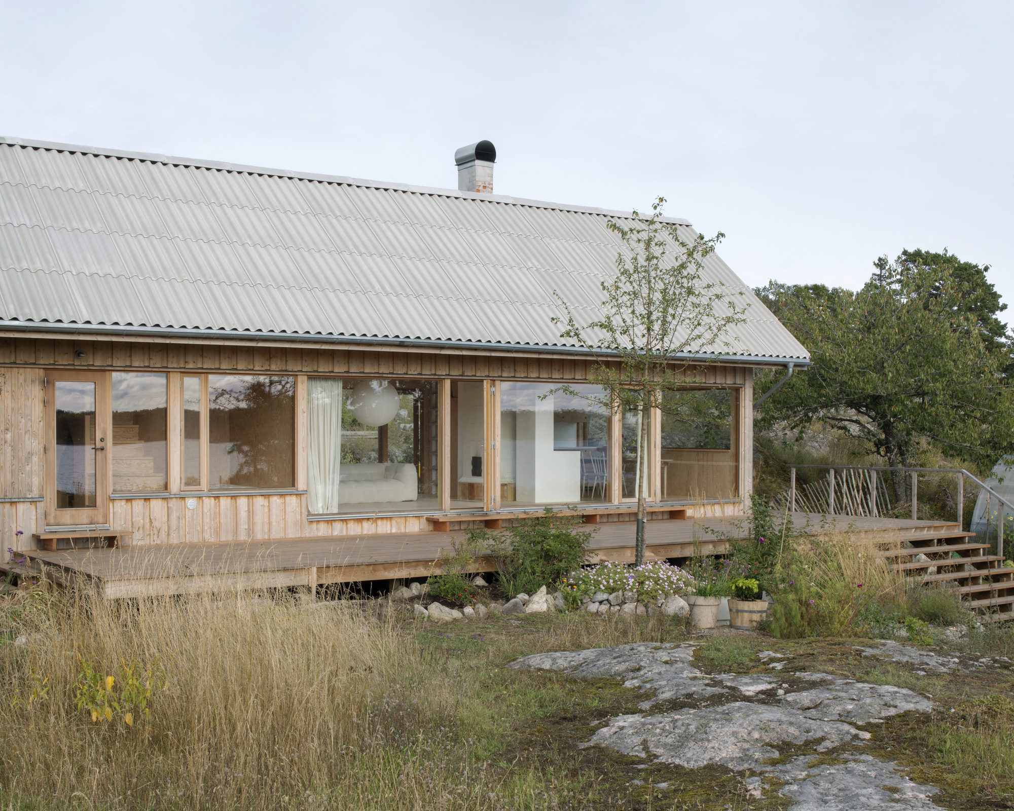 Glazed facade with timber cladding and young tree framed by natural stone and wild grasses