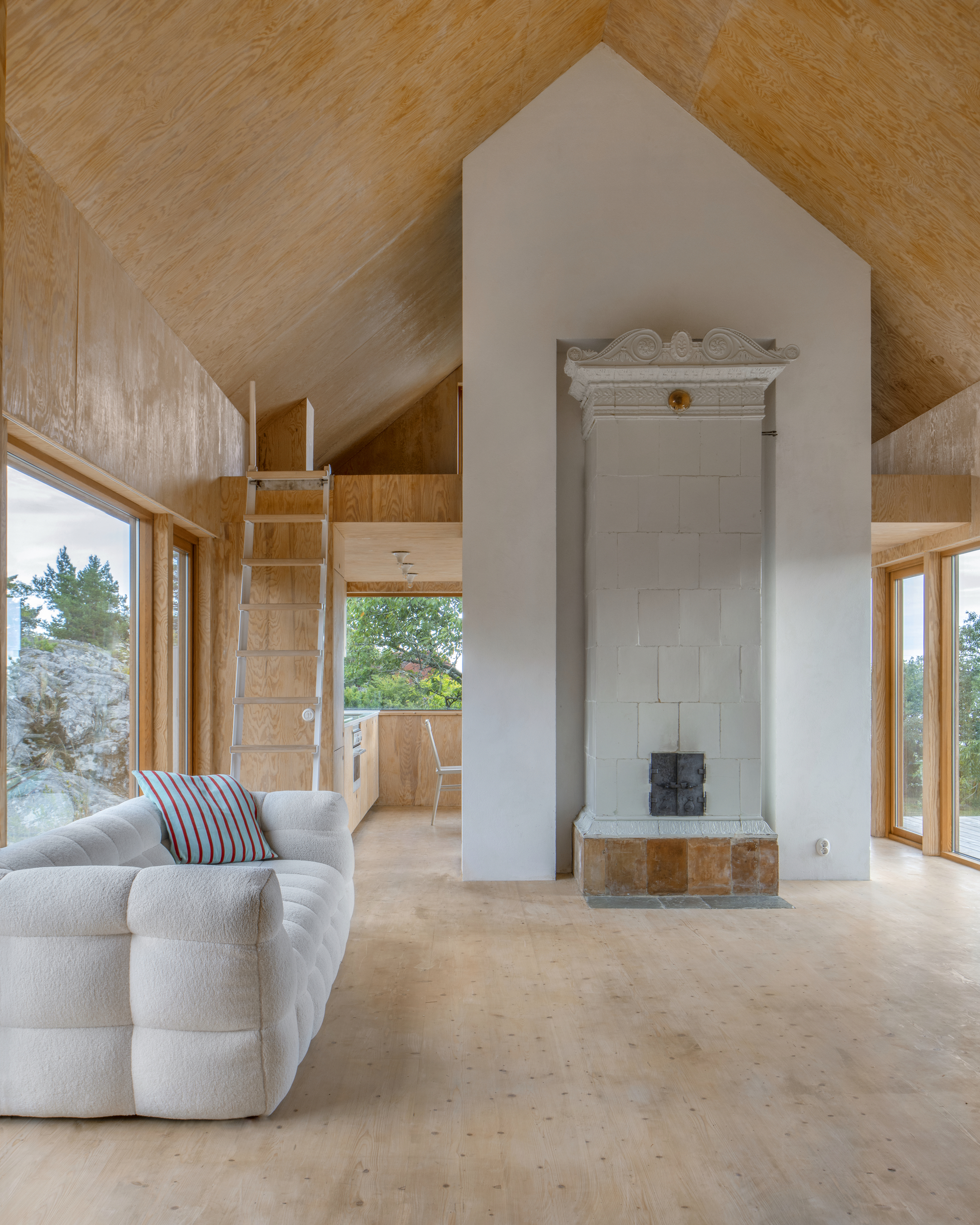 White masonry stove chimney rising through plywood lined interior with vaulted ceiling and loft ladder