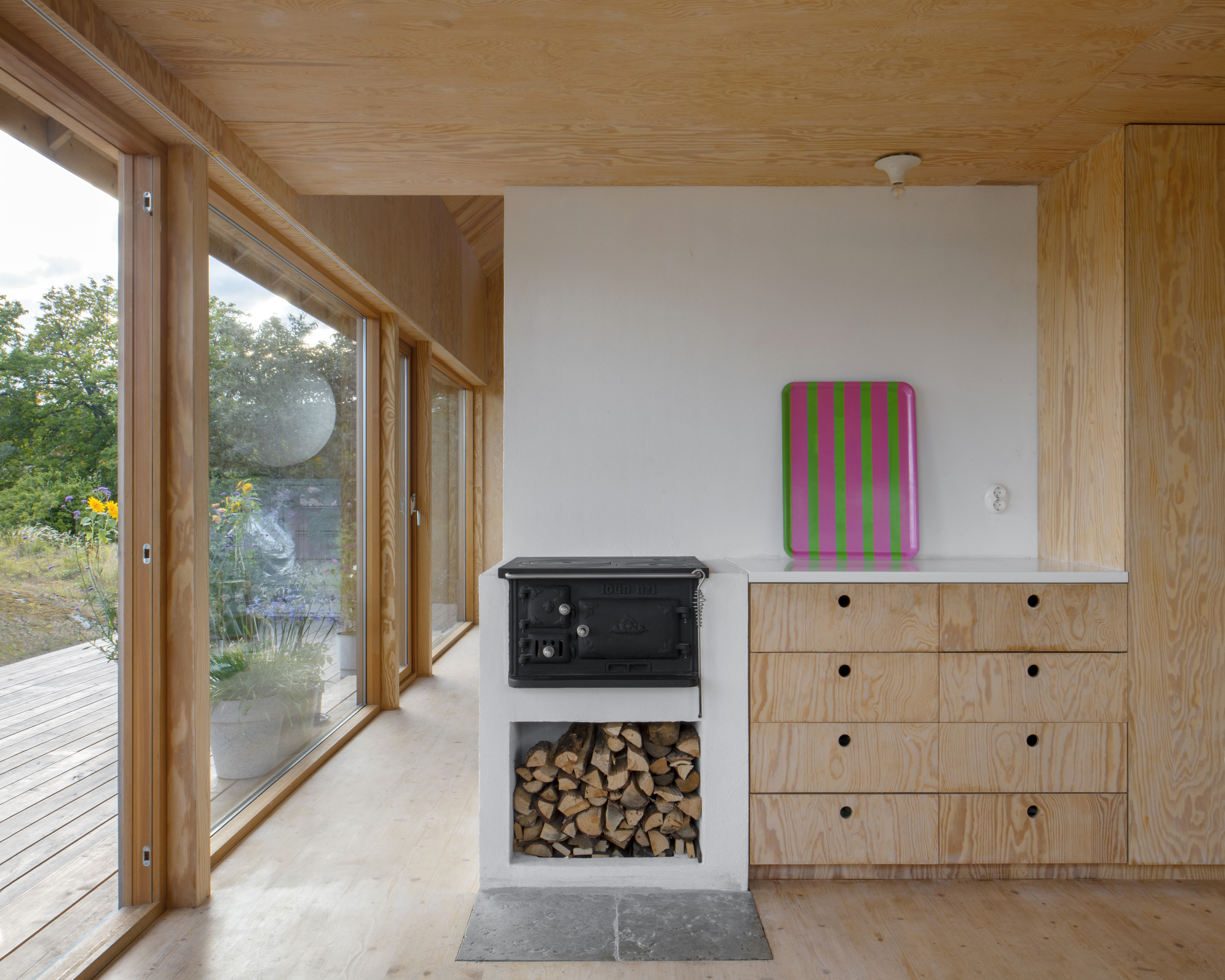 Cast iron wood stove with firewood storage niche beside plywood cabinetry under a timber ceiling