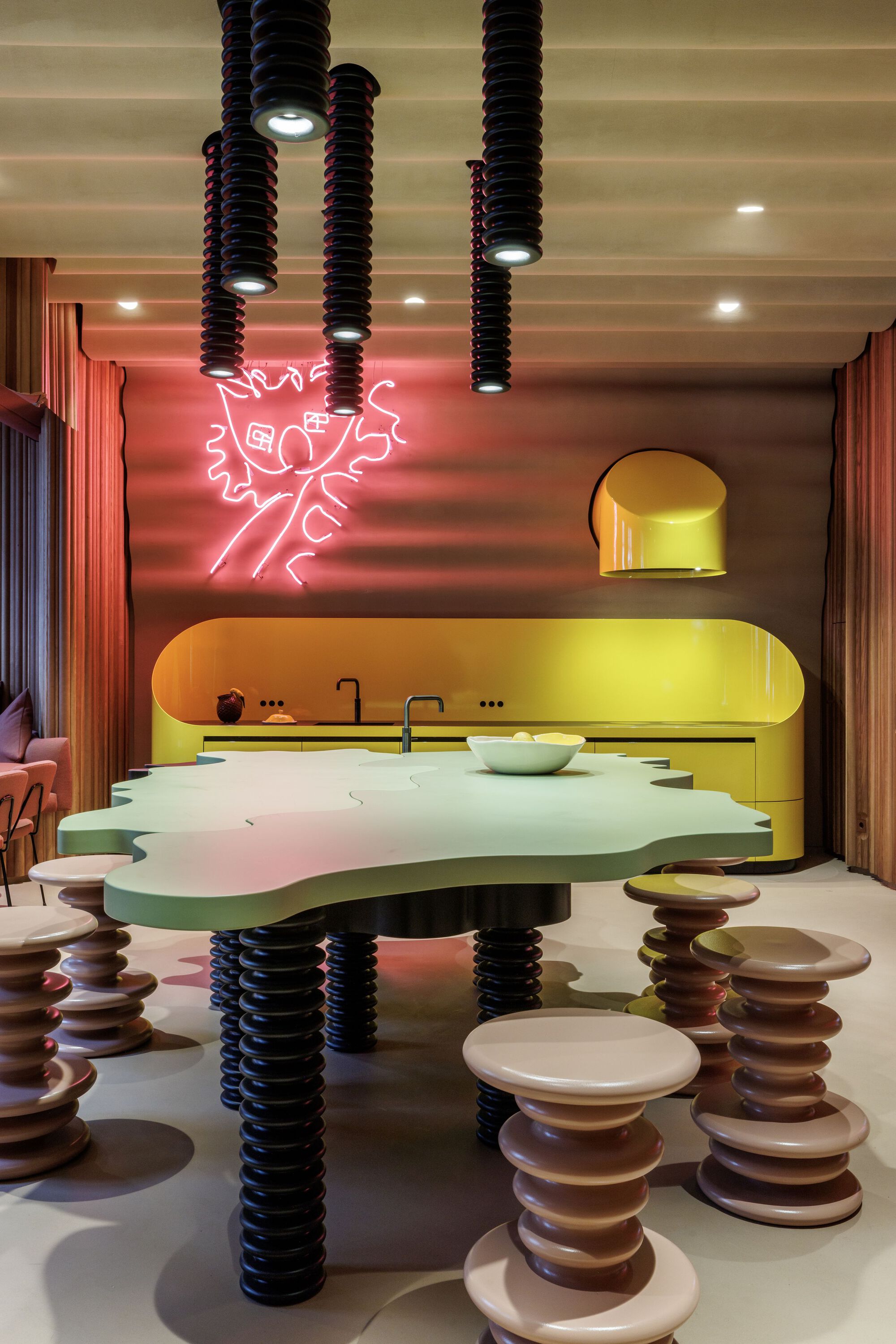 Open kitchen with yellow cabinetry, reflective floor, stacked disc stools, and pink neon wall art