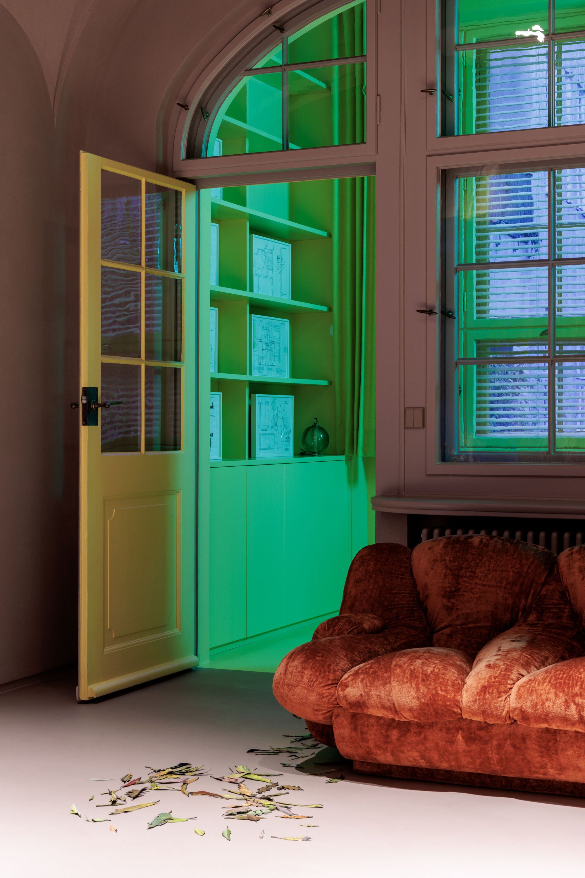 Green-lit alcove with open shelving viewed through yellow framed door beside arched window and velvet sofa