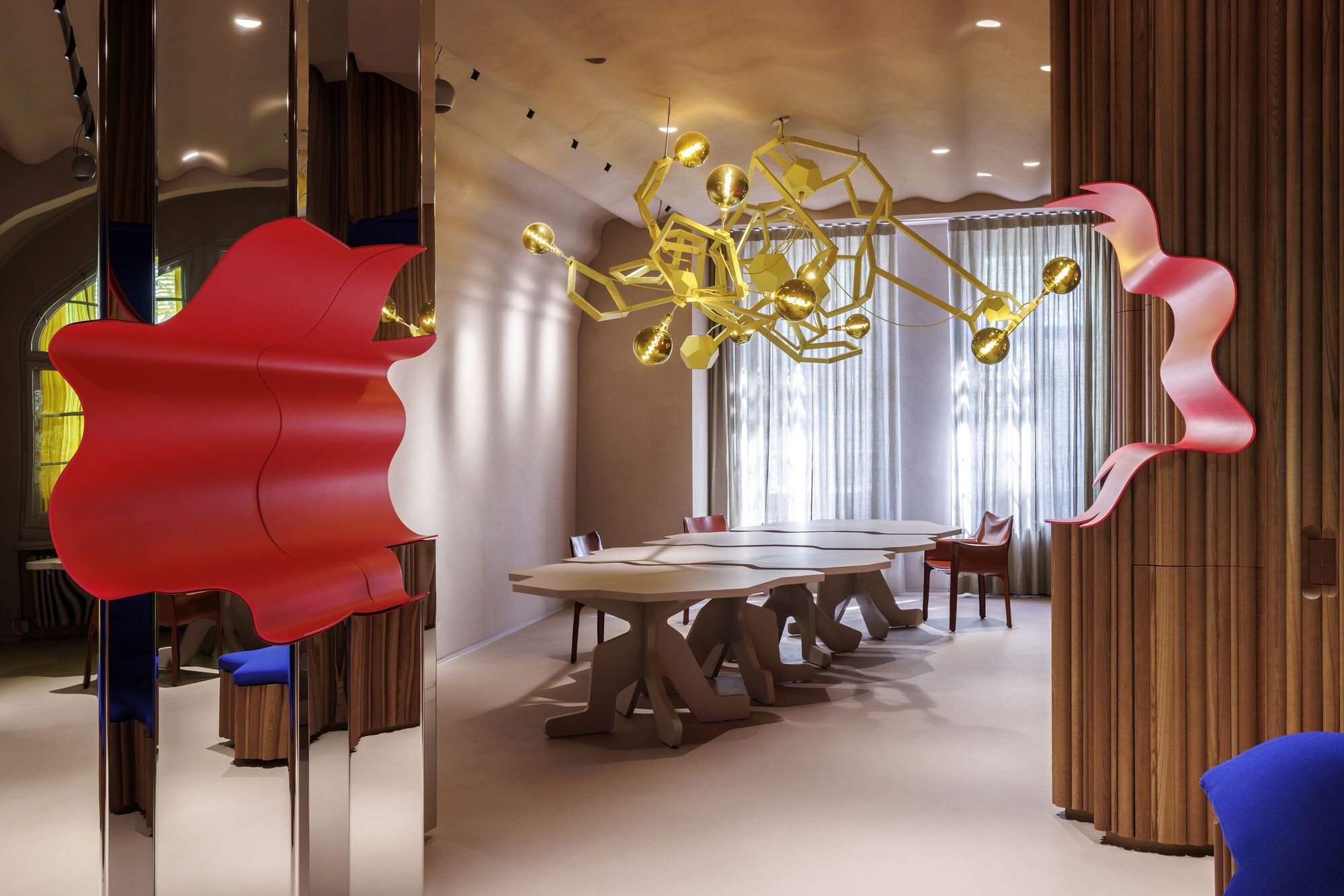 Interior view through red sculptural partition into dining area with gold geometric chandelier above timber table