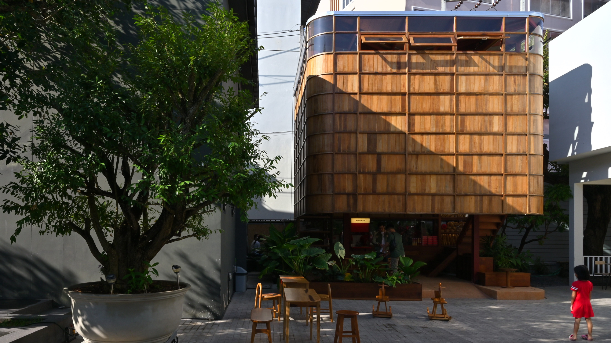 Gridded timber facade of raised volume beside a tree casting shadows across the courtyard below