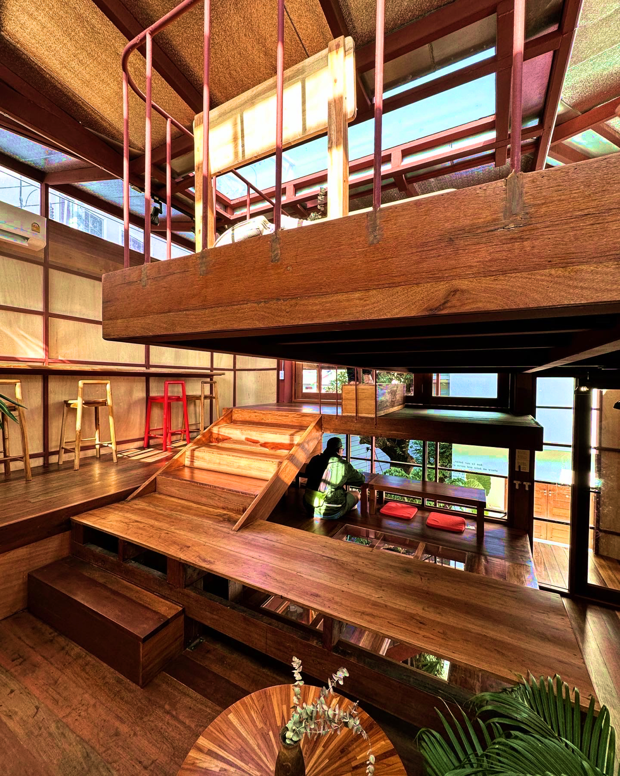 Stacked timber mezzanines and red steel frames seen from below with woven ceiling panels and natural light
