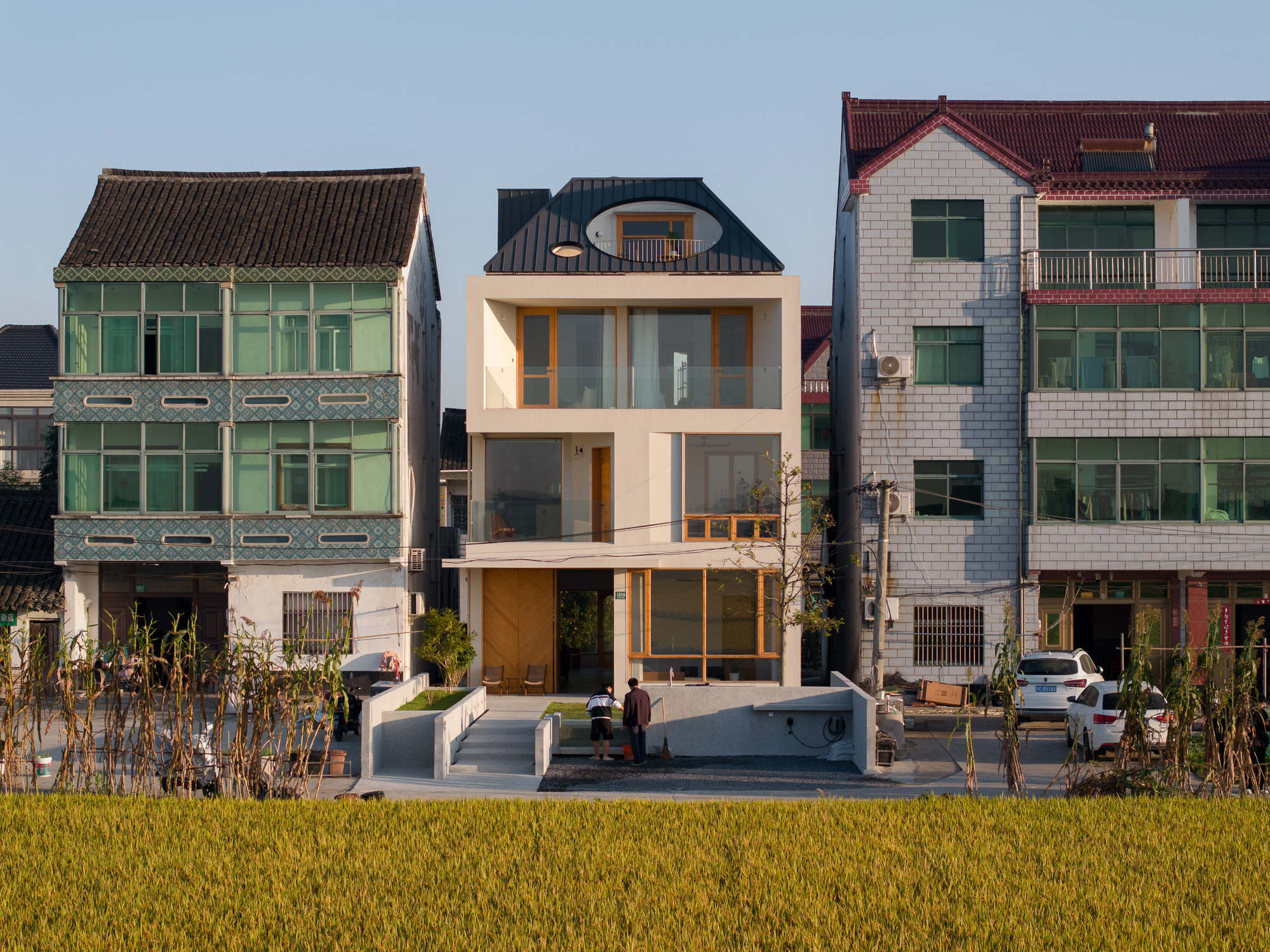 Street facade with white render and timber framed openings standing among older houses and a golden rice paddy