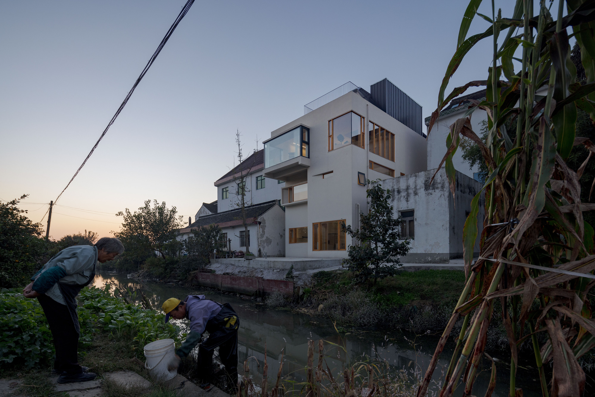 Rear elevation at dusk with residents gathering by the canal edge below the illuminated timber window frames
