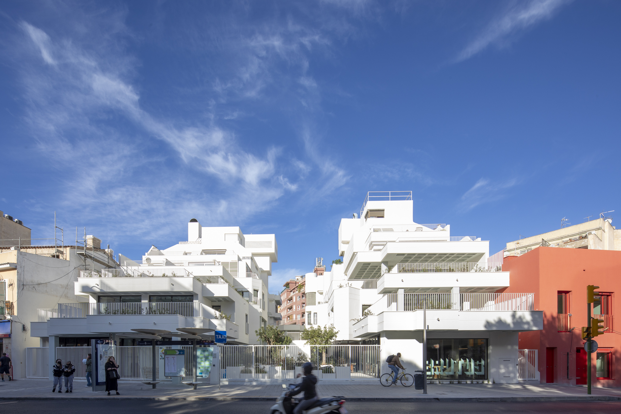 Street view of white stepped housing volumes with vertical louvers and pedestrians below wispy clouds