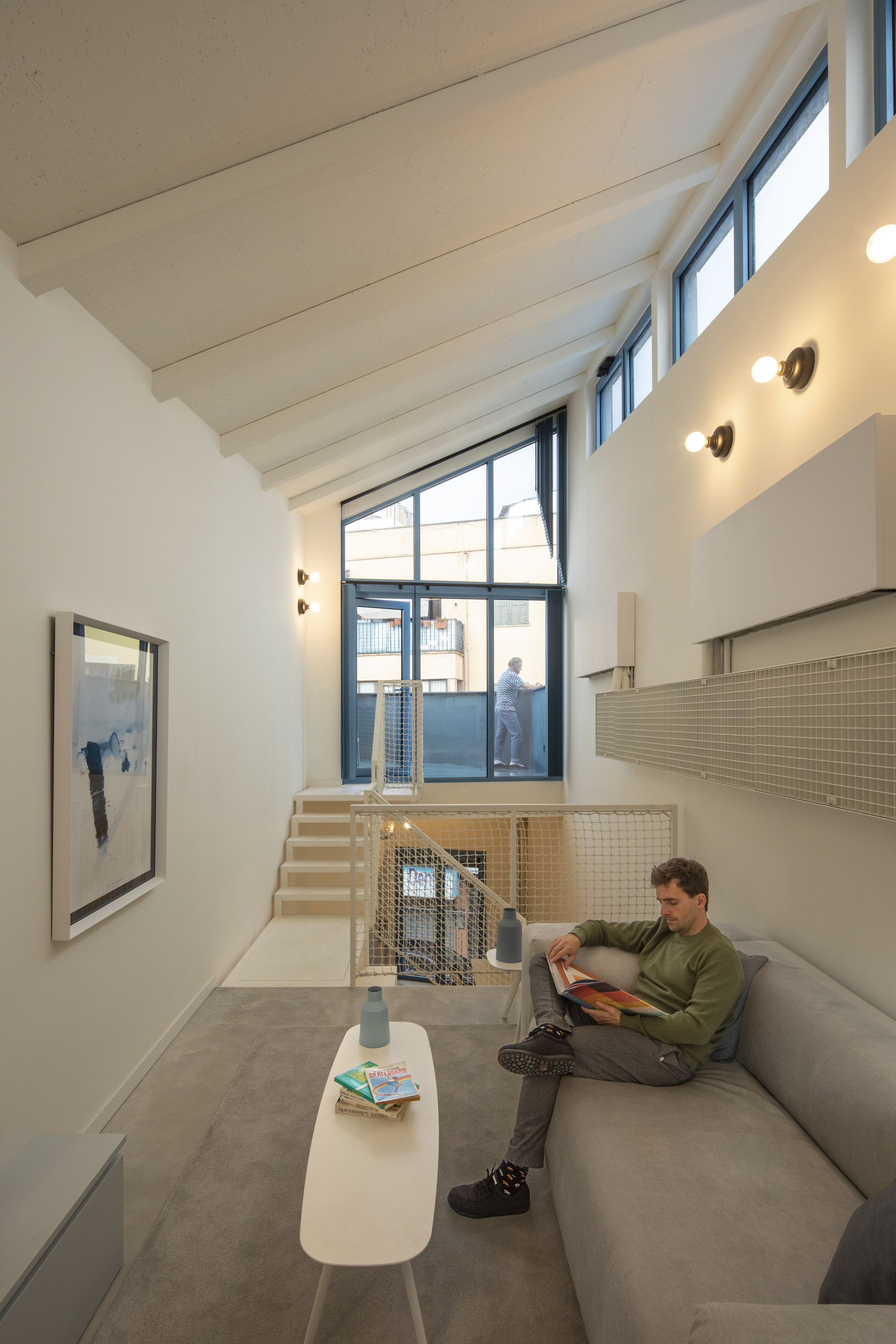 Double-height living room with painted timber ceiling beams and person reading on sofa below mezzanine
