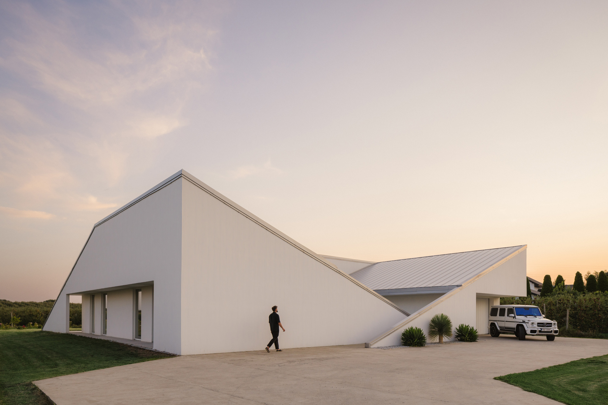 White gabled volume with recessed openings and a figure walking across the paved forecourt at dusk