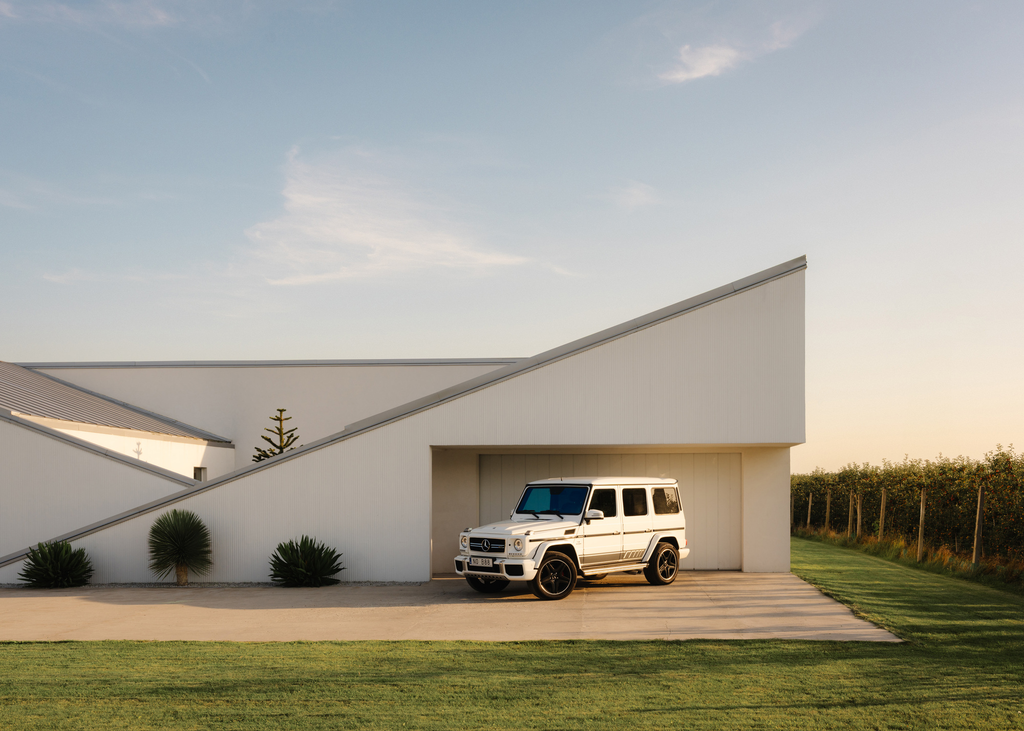 Recessed carport sheltering a white vehicle beneath folded white stucco planes at golden hour