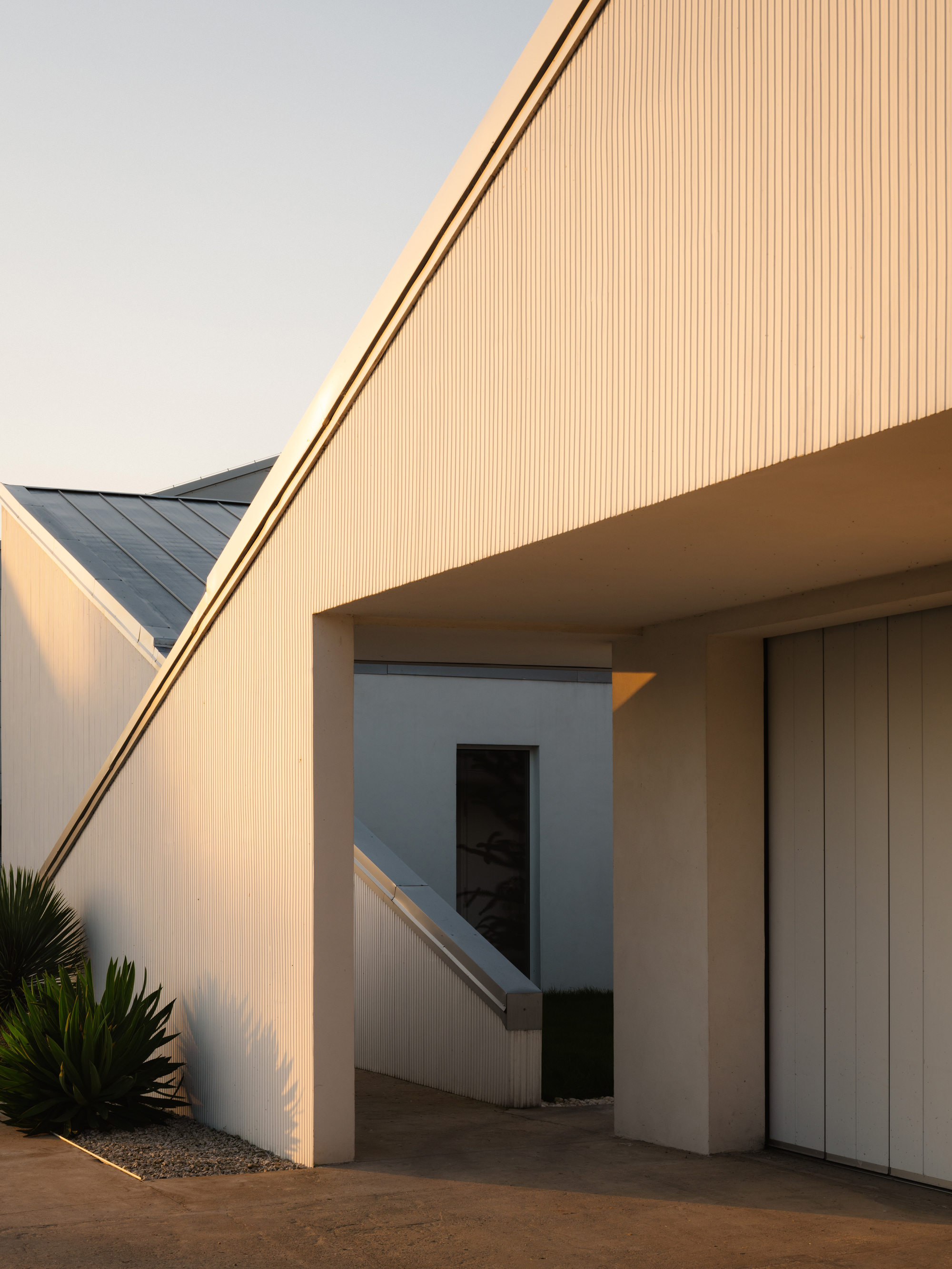 Sloped vertical-ribbed soffit over a courtyard passage at golden hour with planted gravel bed