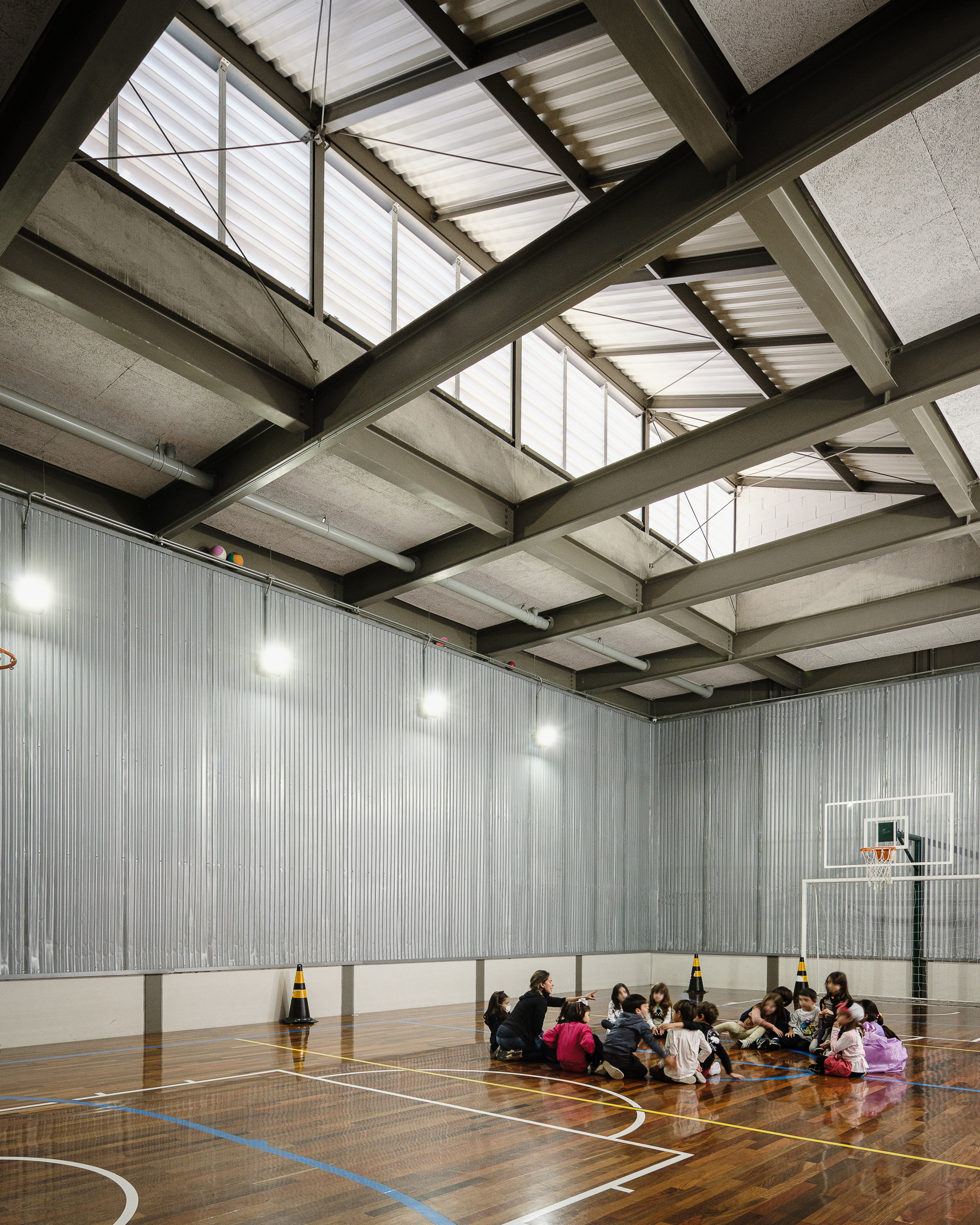 Interior of a gymnasium with exposed concrete beams, triangular skylights, and a group seated on the timber floor