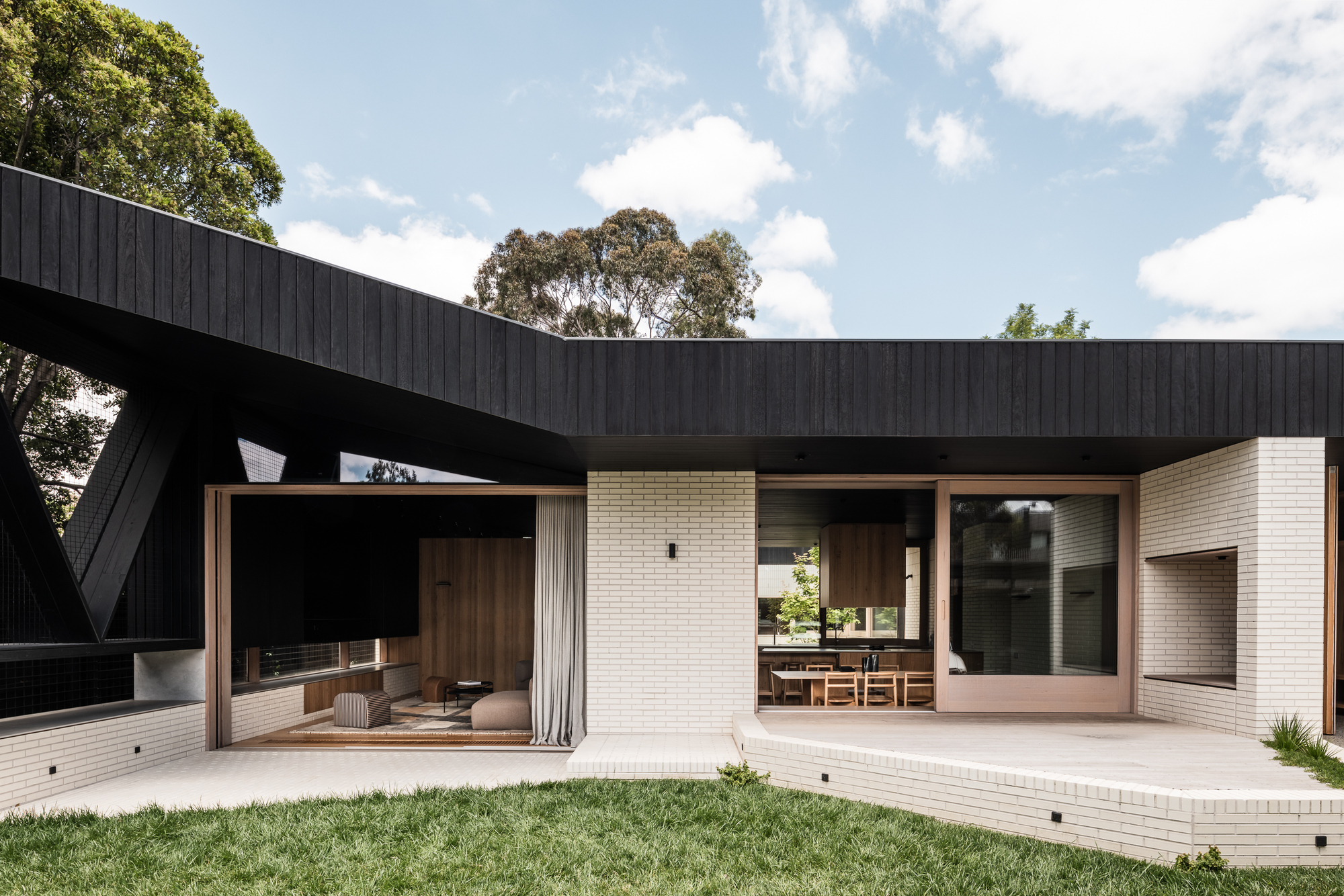 Rear courtyard with white brick walls, timber-framed glazing and a cantilevered black timber roof under midday sun