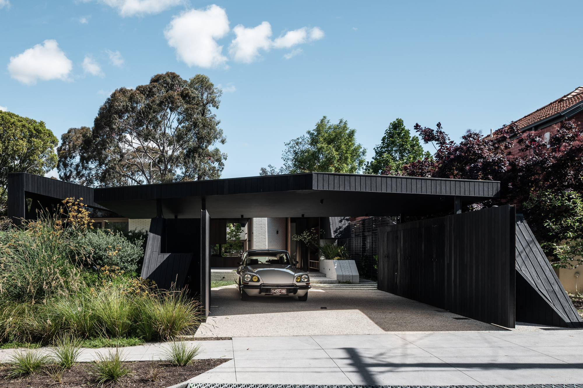 Open carport sheltering a car beneath the projecting roof plane with planted beds on either side