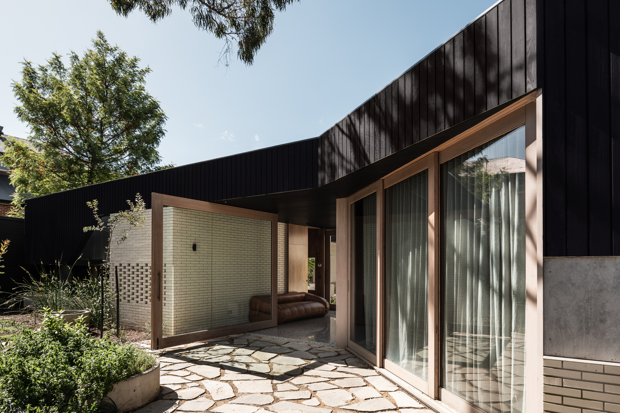 Courtyard with broken flagstone paving, cream brick screening wall and timber-framed glazing under a black timber soffit