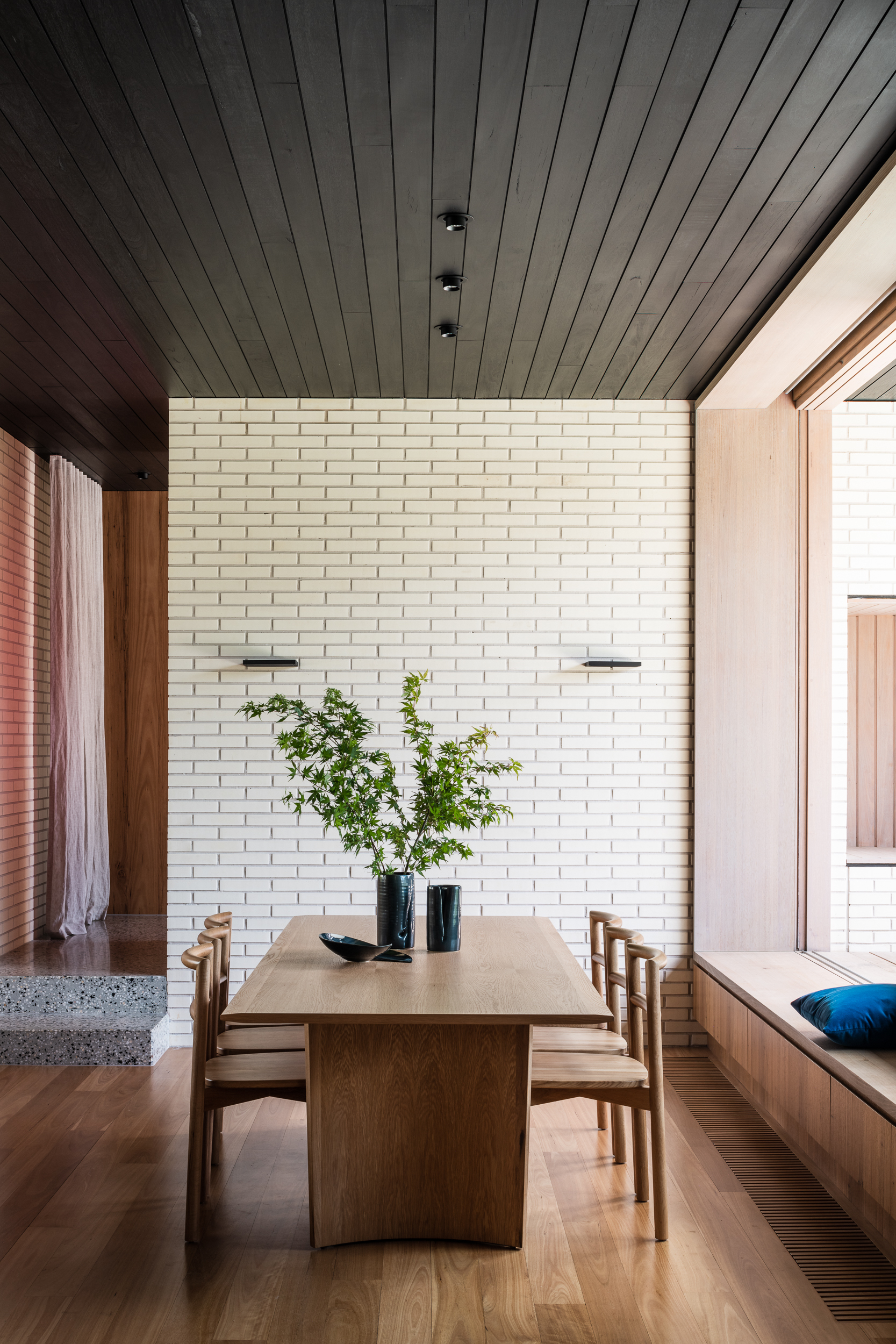 Dining space with timber table and chairs against cream brick wall under black timber ceiling with skylight