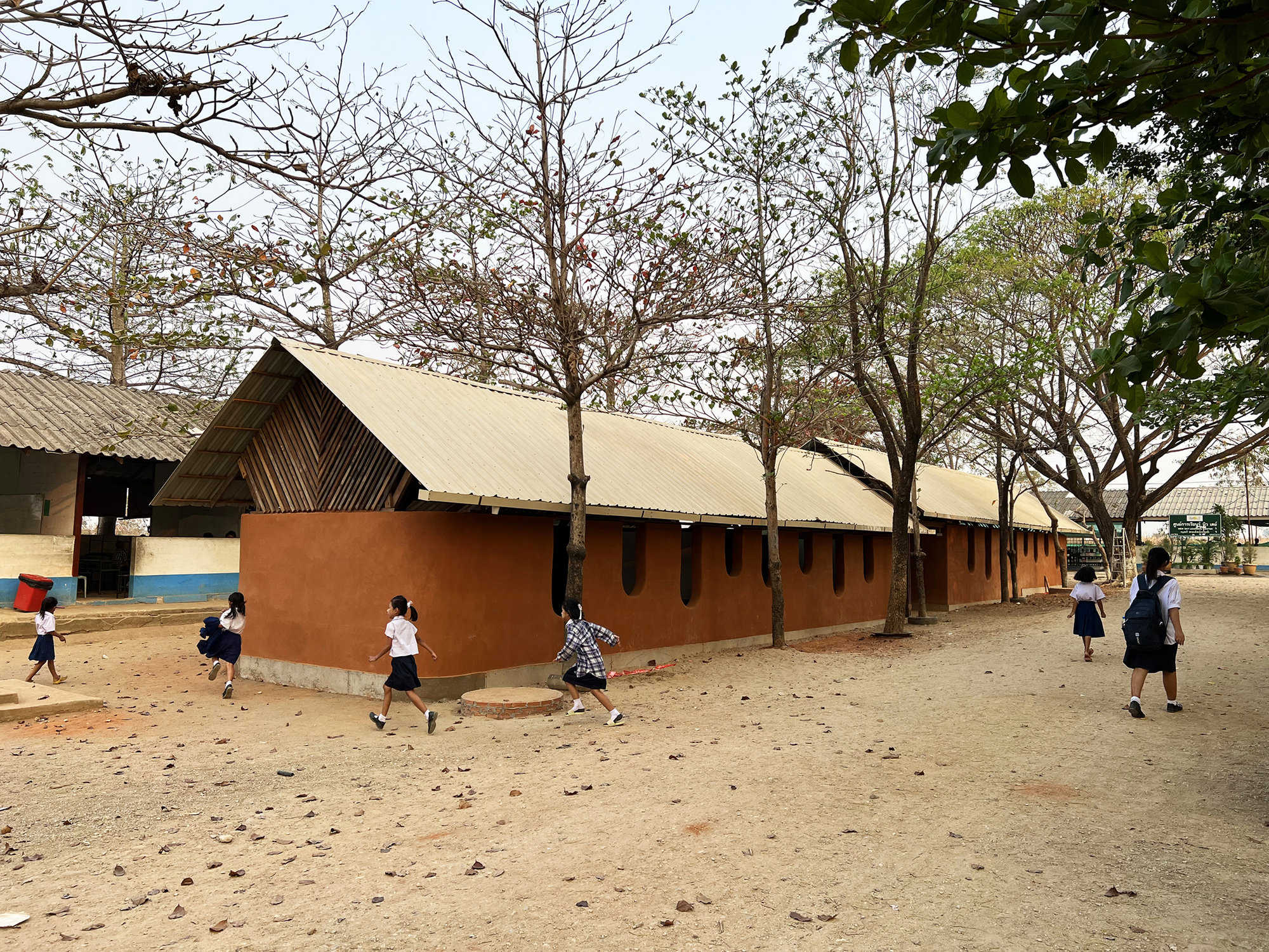 Rammed earth classroom block with corrugated metal gable roof and children running across a sandy courtyard