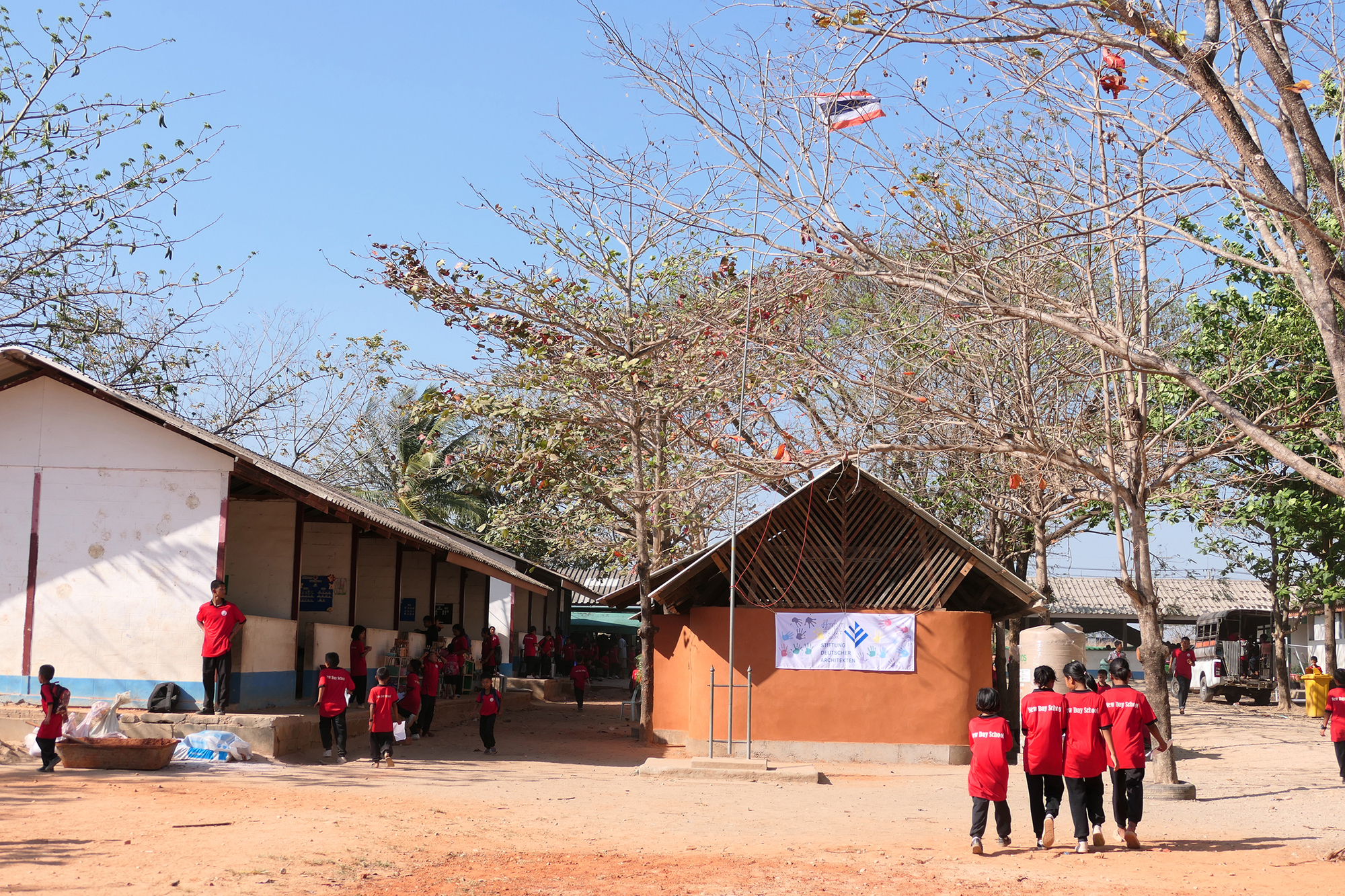 School courtyard showing rammed earth pavilion with pitched roof surrounded by bare deciduous trees and students in red uniforms