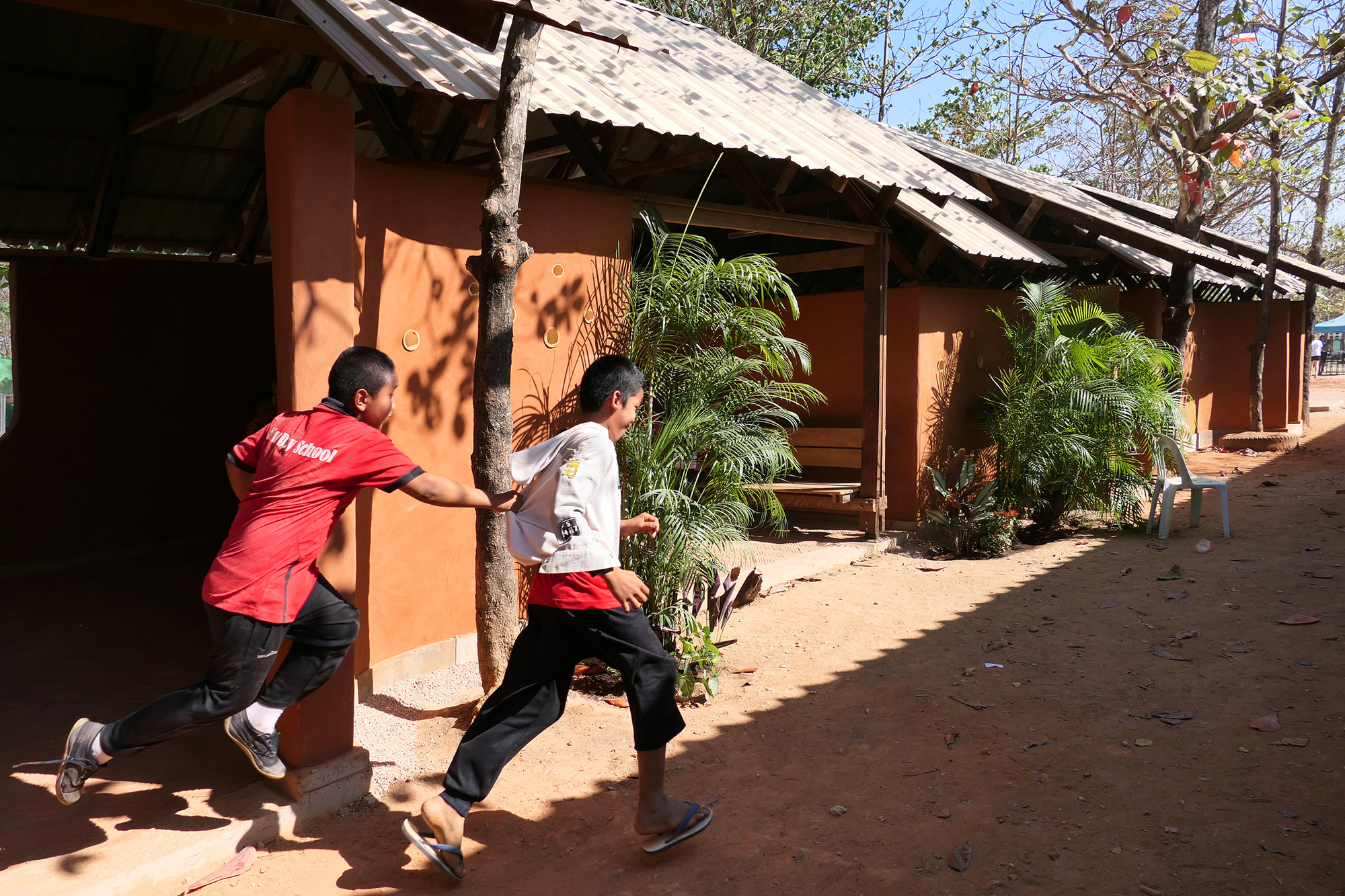 Two children running past the rammed earth facade with planted palms in midday sun