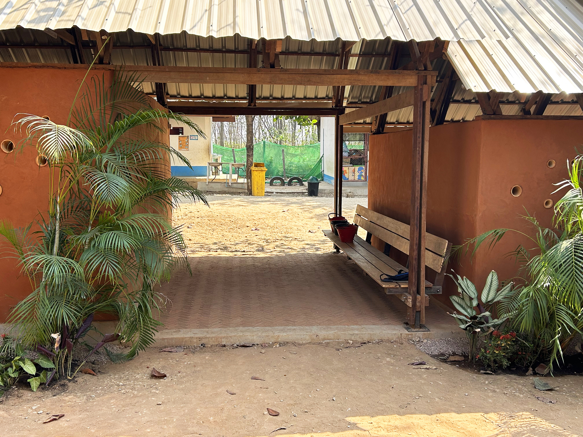 Covered passageway with exposed timber rafters, corrugated metal roof, and potted palms at the entry threshold