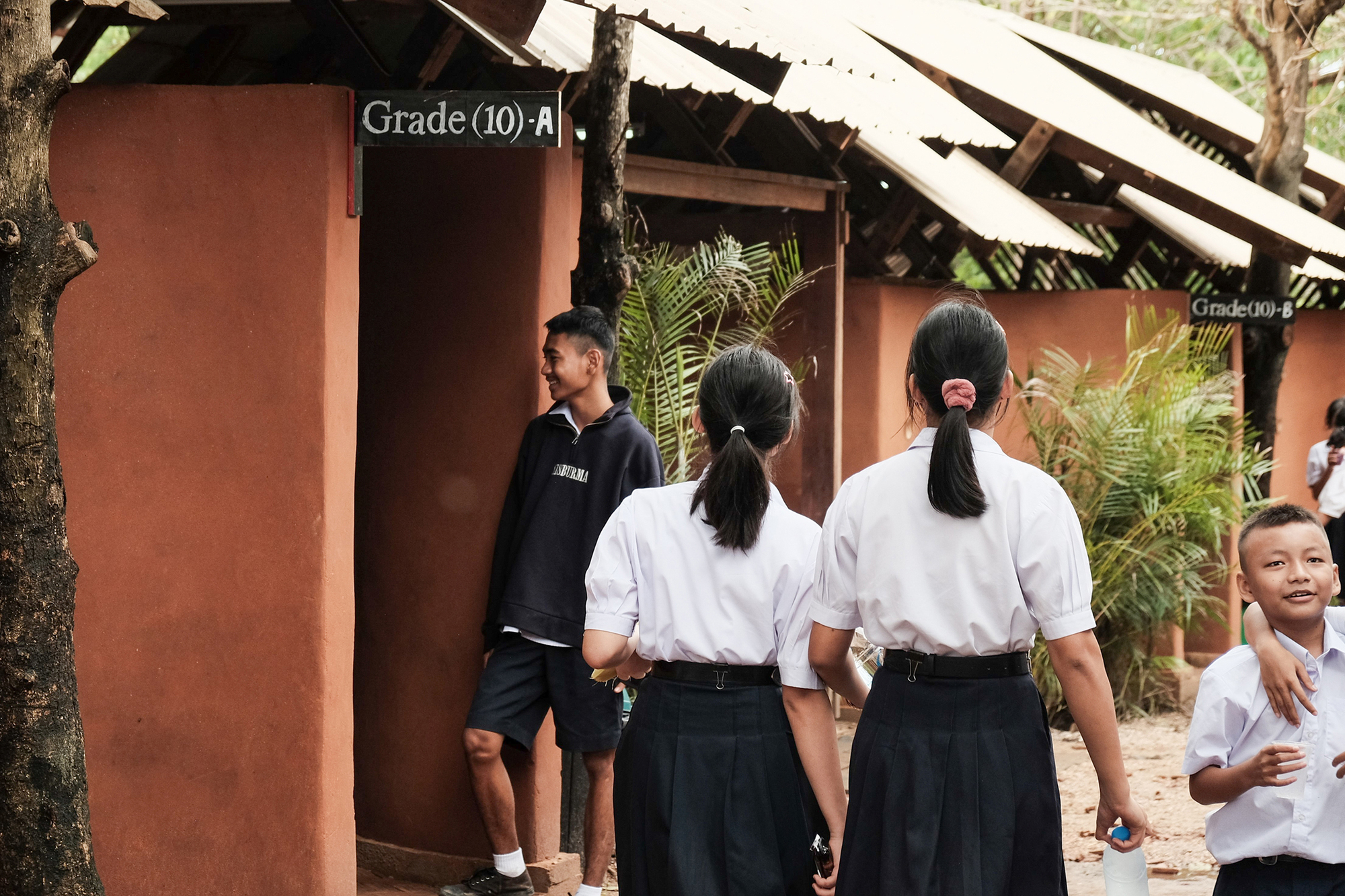 Students gathering outside classroom entries marked with grade numbers under the sloped corrugated roof