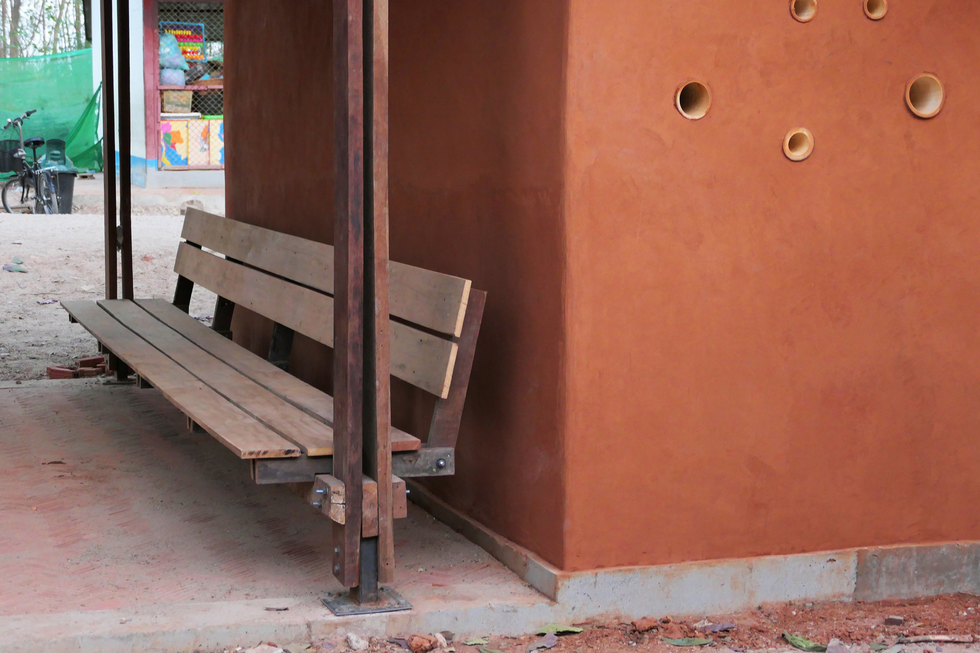 Timber bench against a red ochre wall with circular apertures and steel column