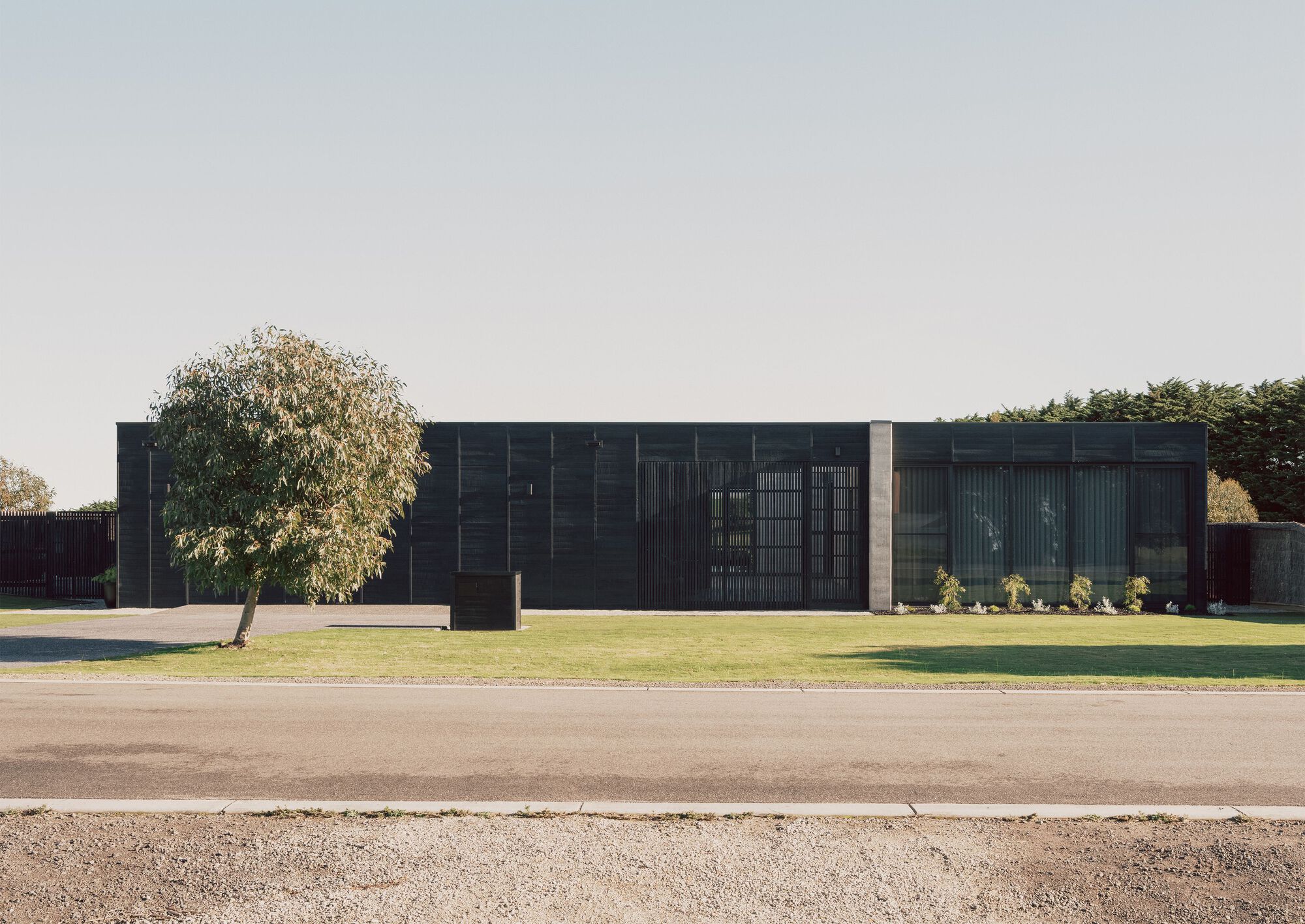 Street view of the dark timber facade with vertical louvres beside a mature tree on the lawn
