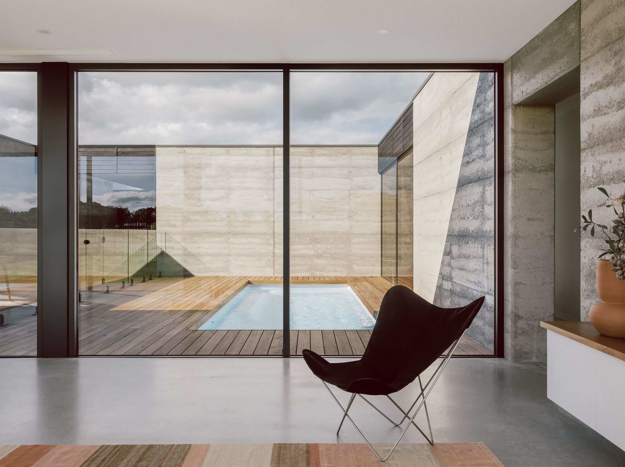 Glass-walled living space overlooking timber deck courtyard with pool and boardformed concrete walls beyond