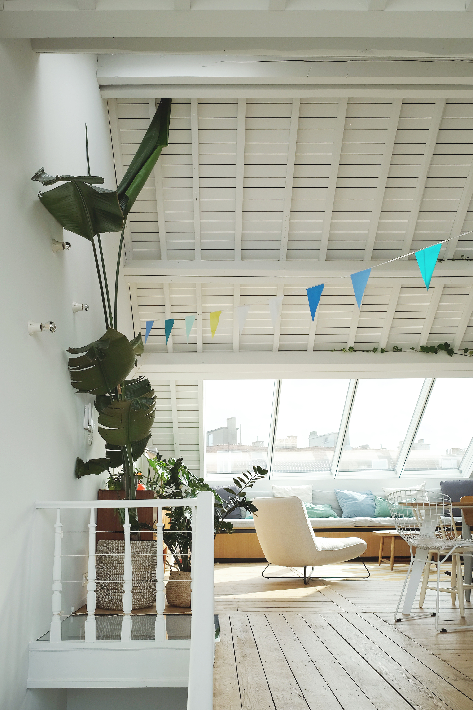 Upper level living space with white beamed ceiling, sloped glazing and potted tropical plants near the stair