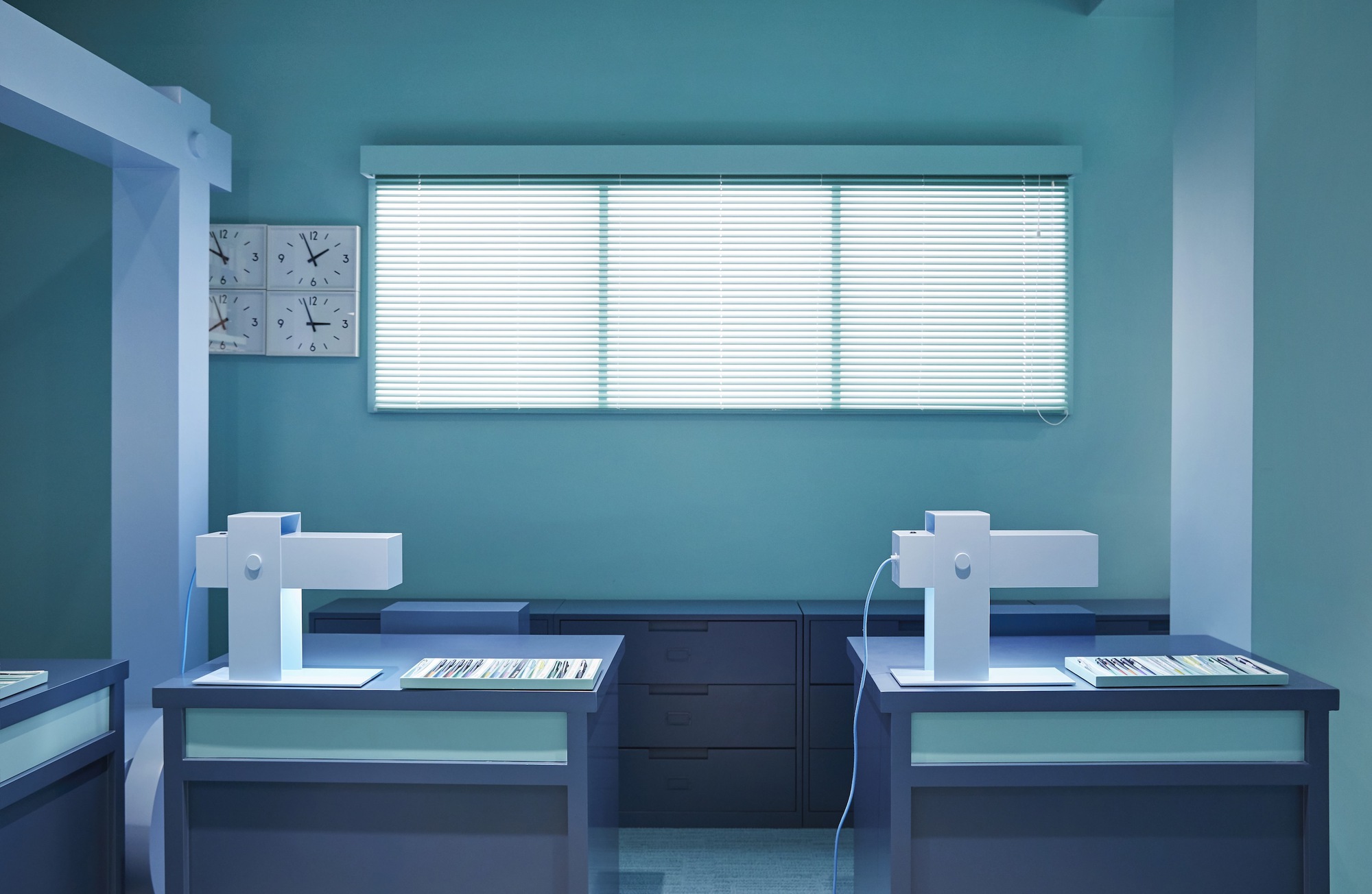 Two mirrored workstations with task lamps beneath horizontal blinds on a turquoise wall with clock grid