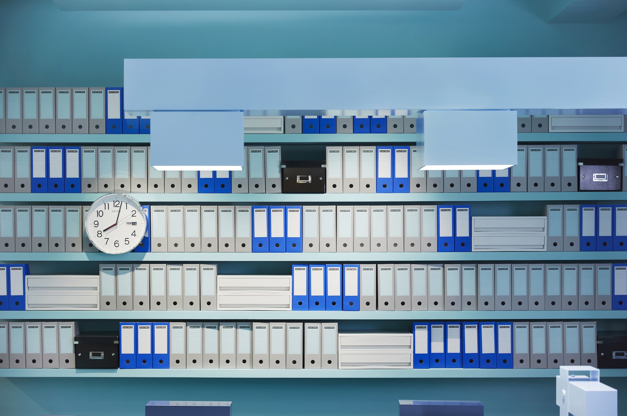 Wall-mounted shelving system displaying rows of blue and grey filing binders with a round clock