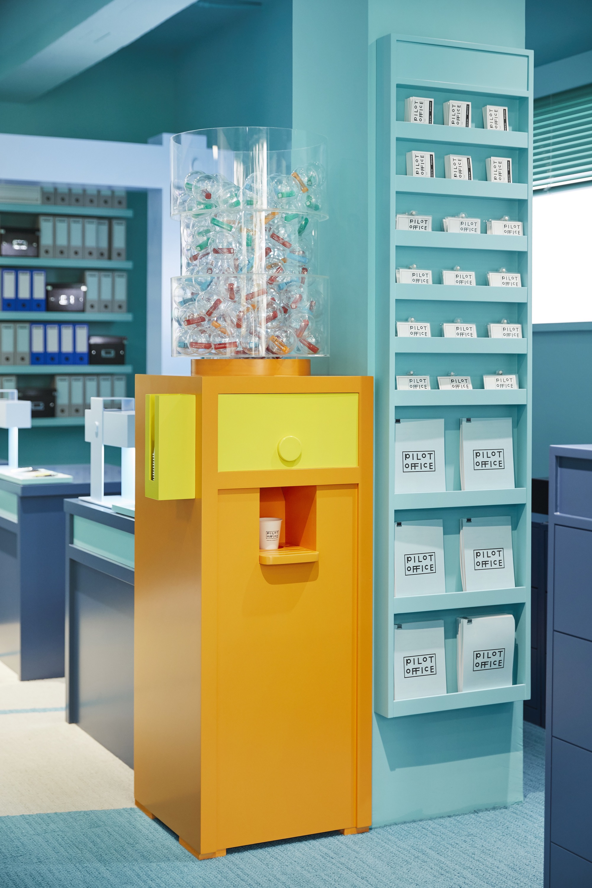 Yellow pedestal dispenser with transparent acrylic top filled with colorful objects beside turquoise shelving unit