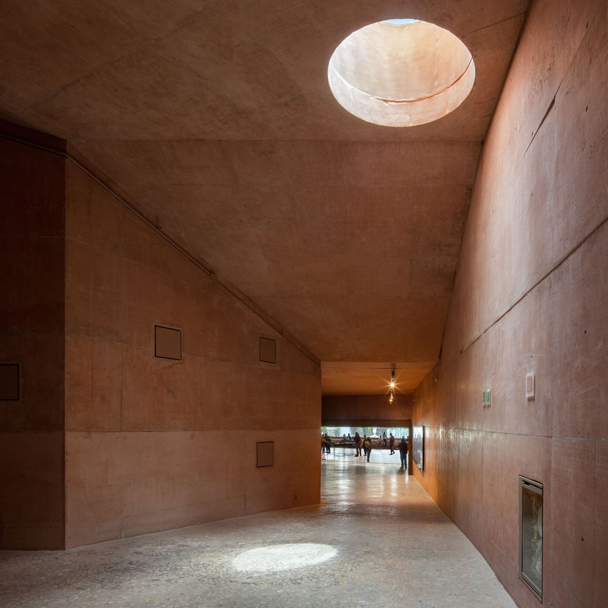 Rammed earth corridor with circular skylight illuminating smooth tapered walls and terrazzo floor