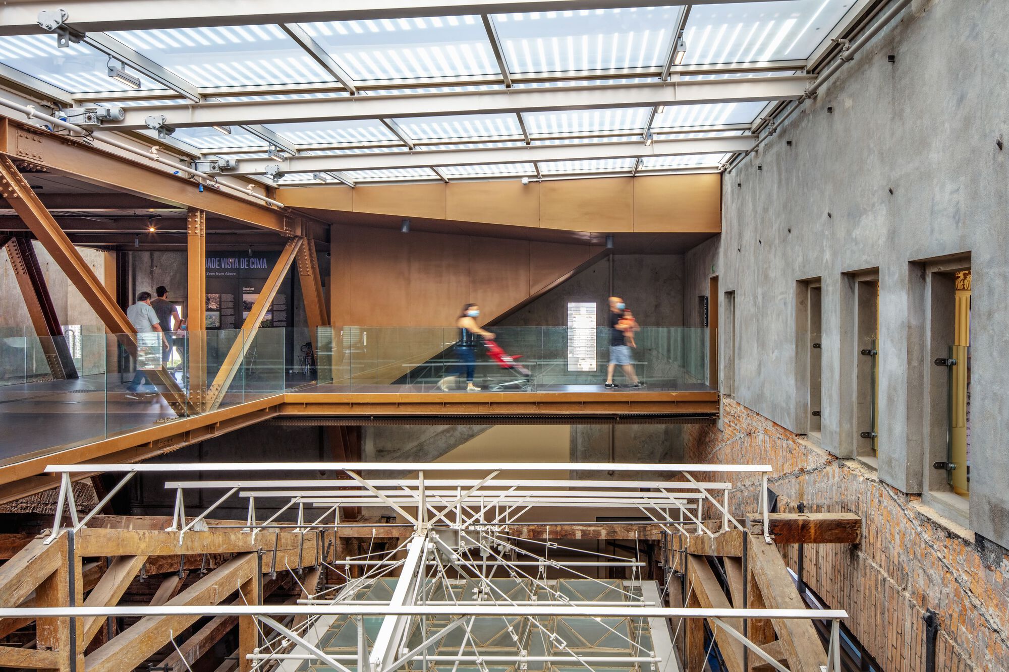 Multi-level atrium with exposed structural steelwork, glass balustrades, and concrete walls under translucent roof