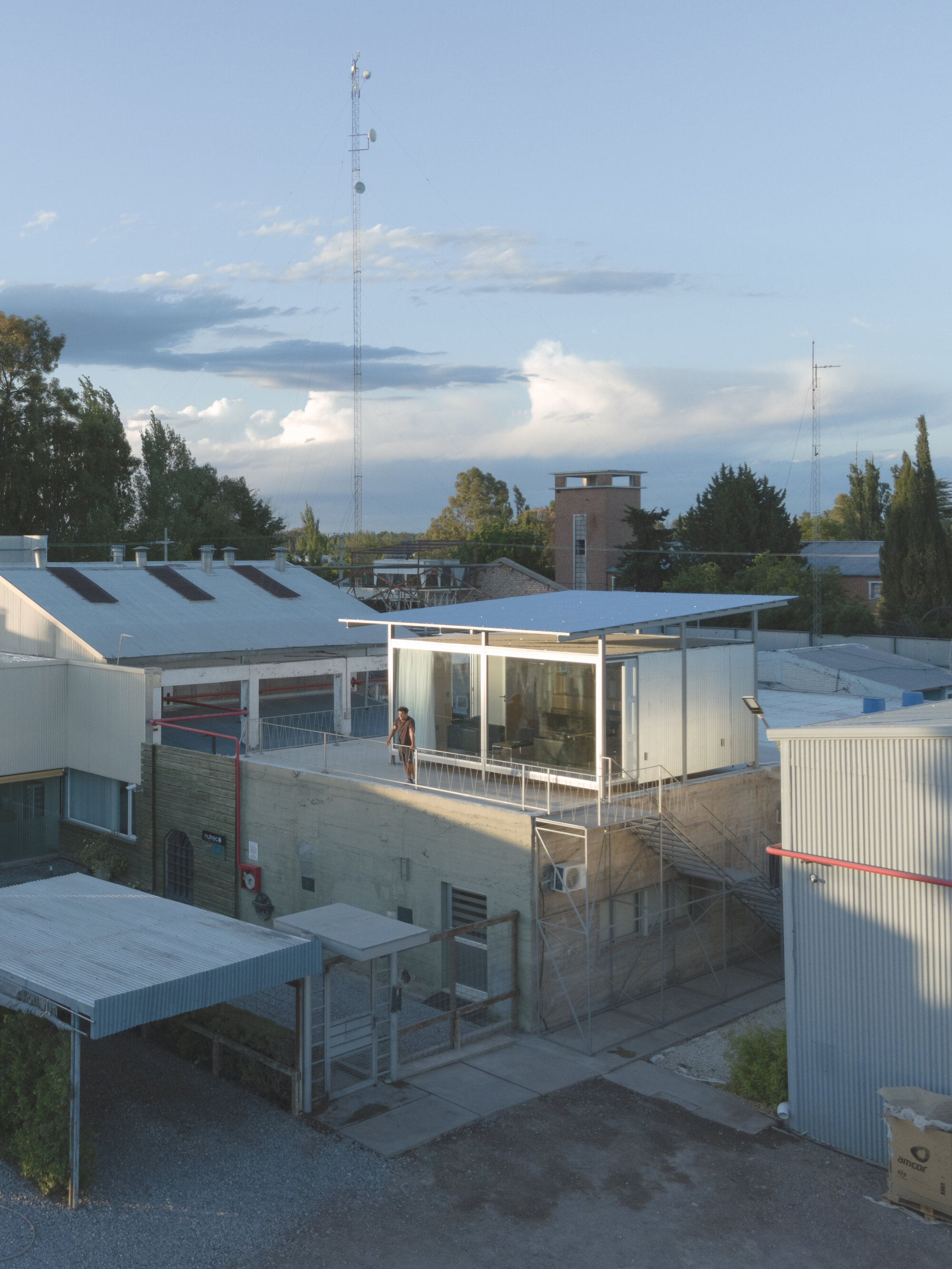 Rooftop pavilion with glazed walls and flat roof perched above industrial buildings at dusk