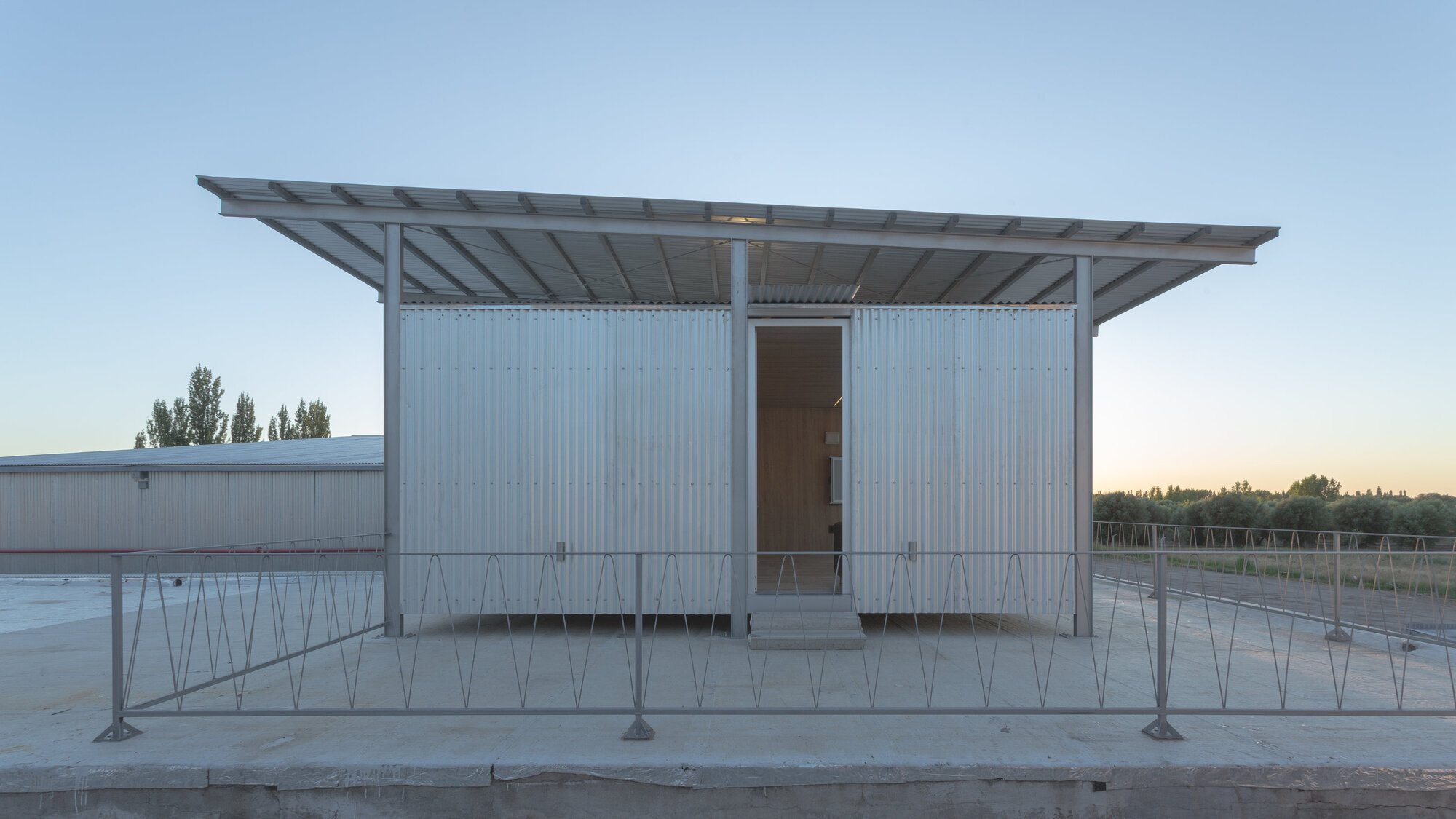 Front elevation of corrugated metal pavilion on a raised platform with exposed steel roof structure at sunset