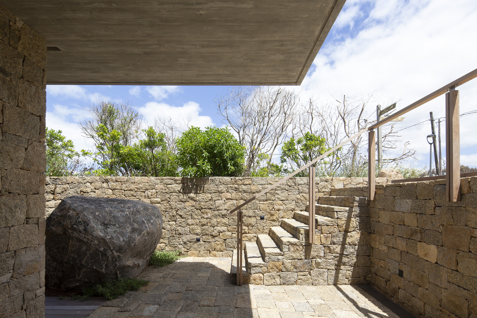 Outdoor courtyard with stone walls, a large boulder, and timber staircase under a concrete soffit