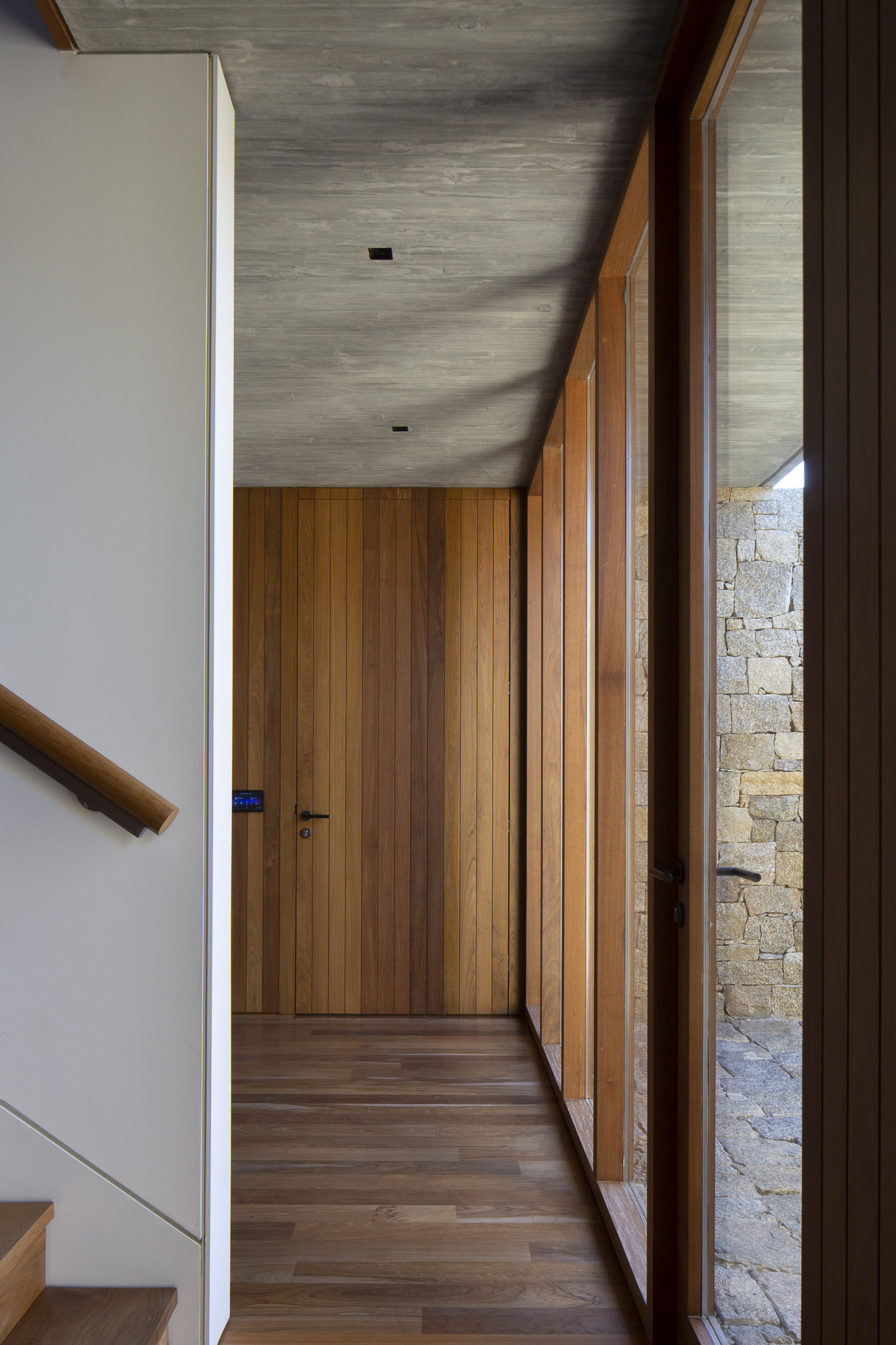 Corridor with board-marked concrete ceiling and timber-lined walls framing a view to the stone wall