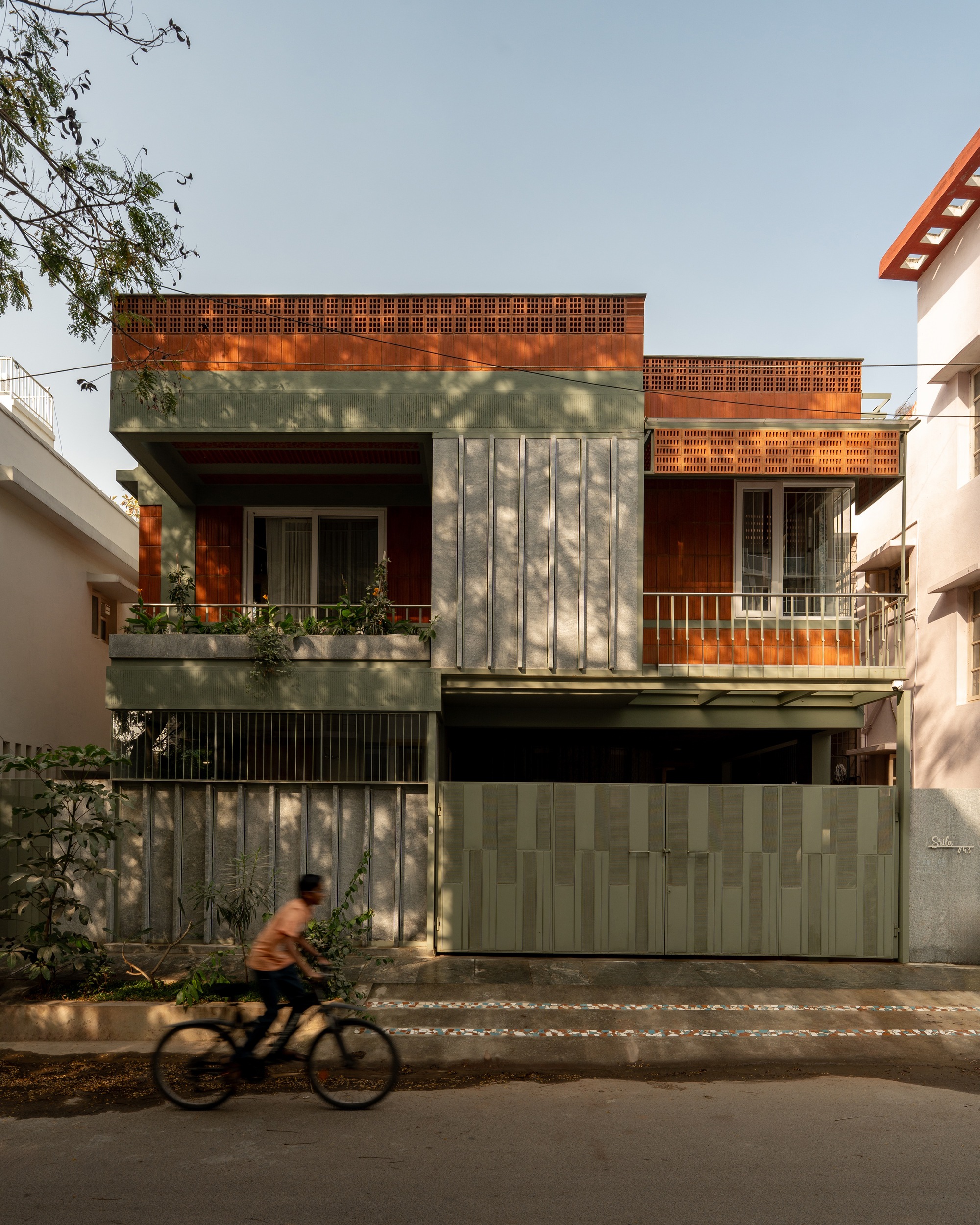 Street facade with vertical concrete panels and perforated brick screens as a cyclist passes in afternoon light