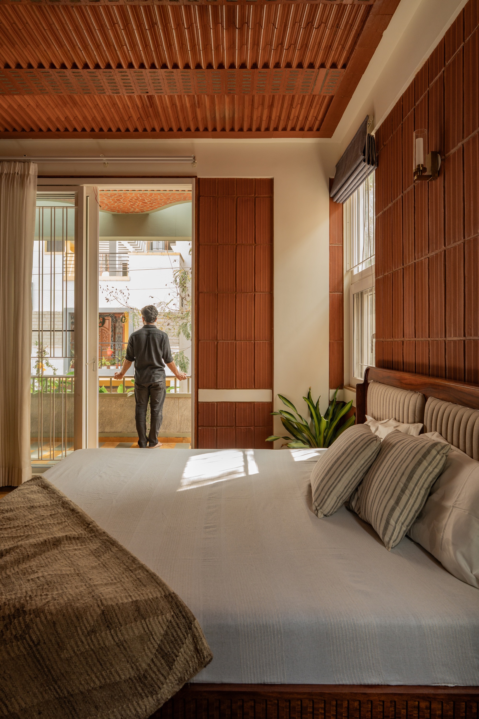 Bedroom with timber-slatted ceiling and wall as a figure opens glazed doors to a sunlit terrace